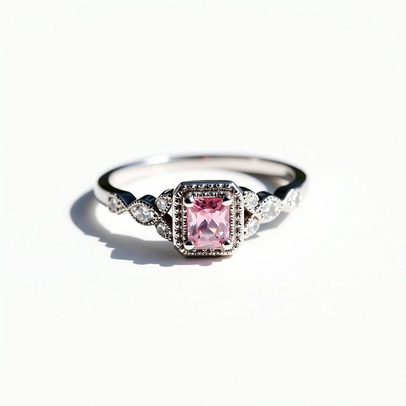 This pink diamond ring features a stunning pink gemstone set prominently at its center in a rectangular cushion cut, surrounded by a halo of smaller diamonds that enhance its brilliance. The band is crafted from a polished metal, likely white gold or platinum, adding a sleek and elegant finish. Delicate diamond accents embellish the shoulders of the ring, set in a marquise pattern that adds a touch of vintage charm. The prong setting holds the central gemstone securely, complemented by the intricate detailing along the band that emphasizes the ring's sophistication and elegance.