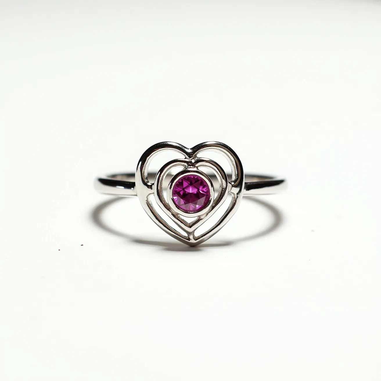 This pink heart ring features a delicate design with a prominent pink gemstone at its center. The gemstone is likely a round cut, set within a bezel to provide a secure and polished look. The metal appears to be a shiny silver, possibly sterling silver or white gold, and is shaped into a double heart motif, adding to its romantic appeal. The ring band itself is sleek and smooth, complementing the heart-shaped centerpiece. The overall design emphasizes elegance and simplicity, making it an attractive accessory for various occasions.