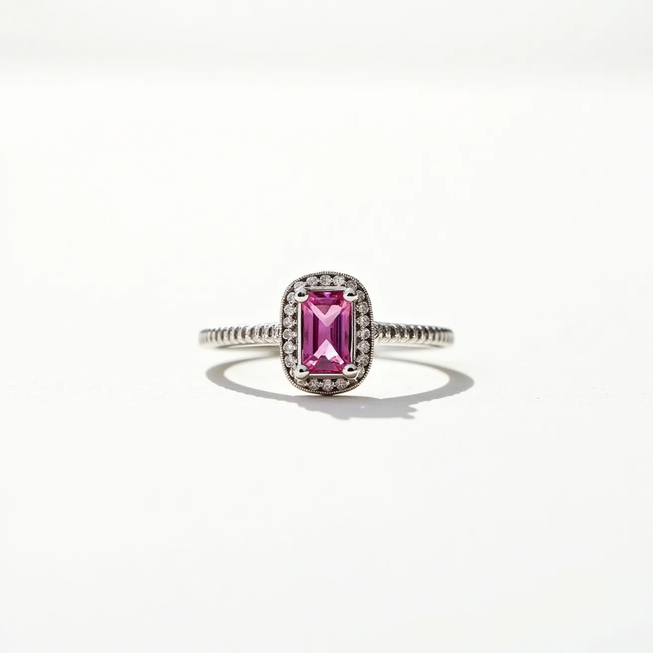 This pink sapphire ring features a striking, rectangular-cut pink sapphire at its center, exuding elegance and vibrancy. The sapphire is enclosed within a delicate halo of small, round-cut diamonds, enhancing its brilliance and adding a layer of intricate detail. The setting appears to be crafted in a polished metal, likely white gold or platinum, which complements the overall aesthetic and provides a secure and sophisticated contrast. The band is intricately designed, potentially with a twisted or rope pattern, adding a touch of uniqueness and refinement to the piece. This ring is both elegant and eye-catching, making it a beautiful accessory for any occasion.