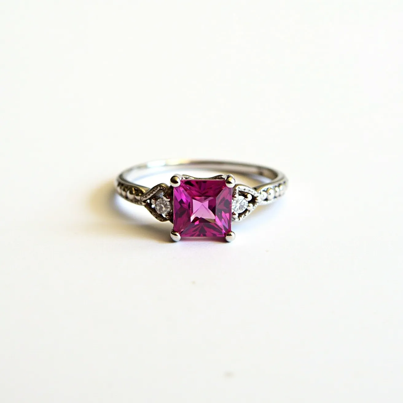 This pink sapphire ring features a striking square-cut pink sapphire as the centerpiece, set in a polished silver or white gold band. The central stone is secured by a four-prong setting, which elegantly elevates it above the band. Flanking the sapphire are two smaller round diamonds on each side, further accentuated with a pave setting that leads into the ring's shank. The combination of the bright pink sapphire and sparkling diamonds creates a visually captivating design, blending classic elegance with a modern touch.