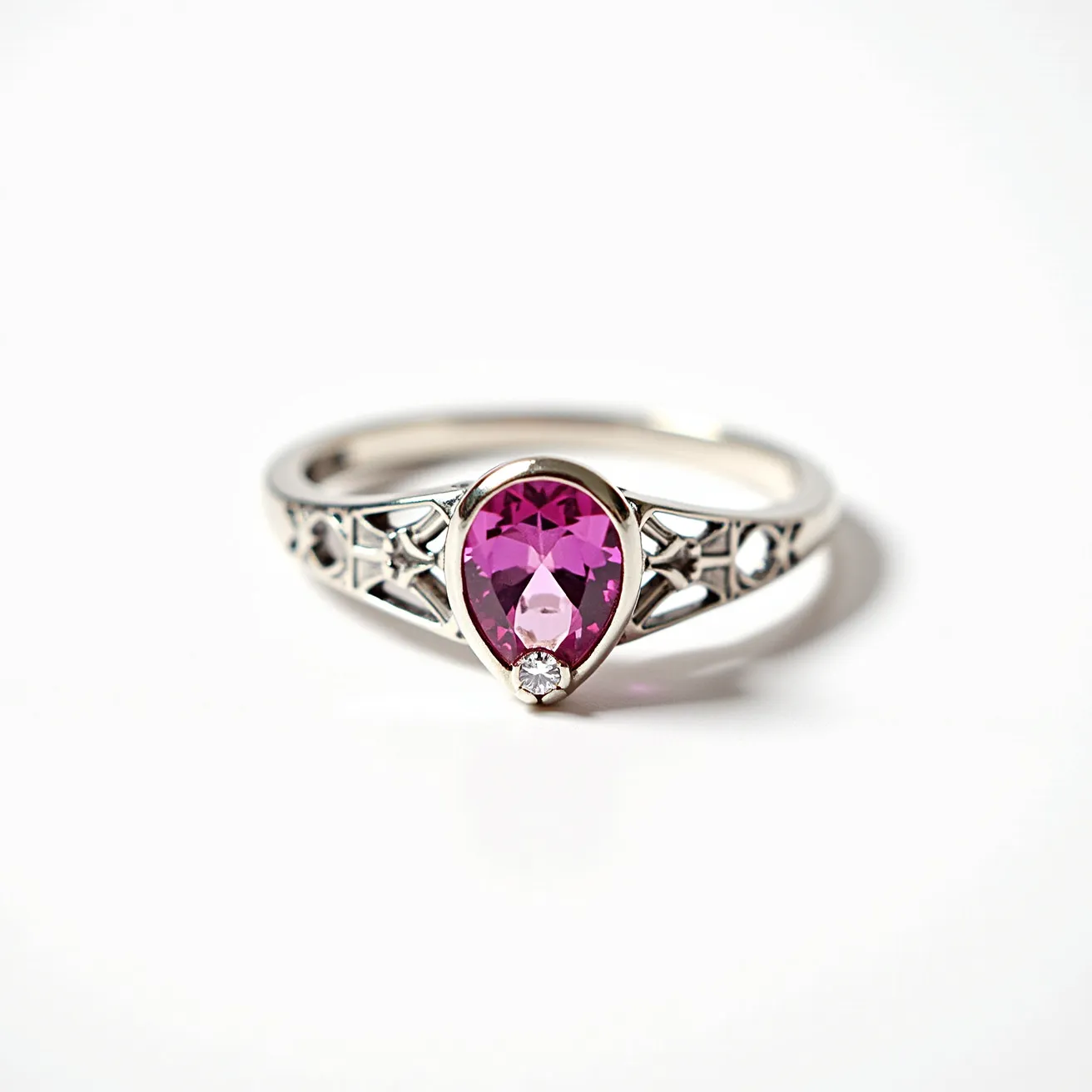 This pink sapphire ring features a stunning pear-shaped pink sapphire, set in a polished, reflective metal band, likely made of white gold or platinum. The sapphire is complemented by a round diamond accent positioned just below the tip of the pear shape, enhancing its brilliance. The band itself is intricately designed with decorative cut-out patterns, adding an elegant touch to the overall design. The sapphire is securely held in a bezel setting, ensuring durability and style.