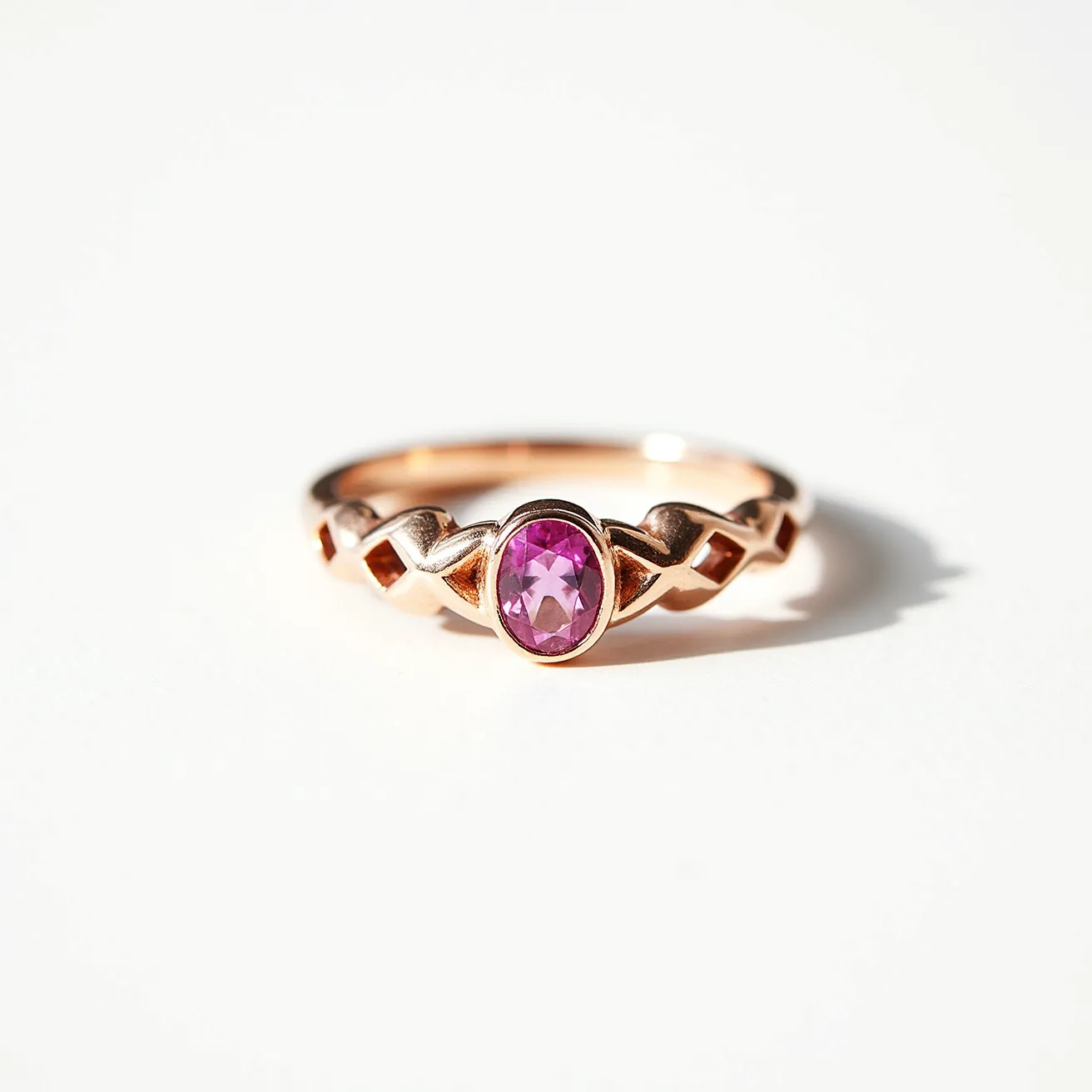 This pink sapphire ring features an alluring oval-cut pink sapphire, faceted to enhance its brilliance and set in a bezel that highlights its vibrant hue. The band appears to be made of rose gold, enhancing the warmth and elegance of the overall design. The band incorporates an intricate open-weave pattern that adds a touch of sophistication and modernity. The seamless integration of the stone with the metal exemplifies a refined craftsmanship, indicative of a carefully considered and executed jewelry piece.