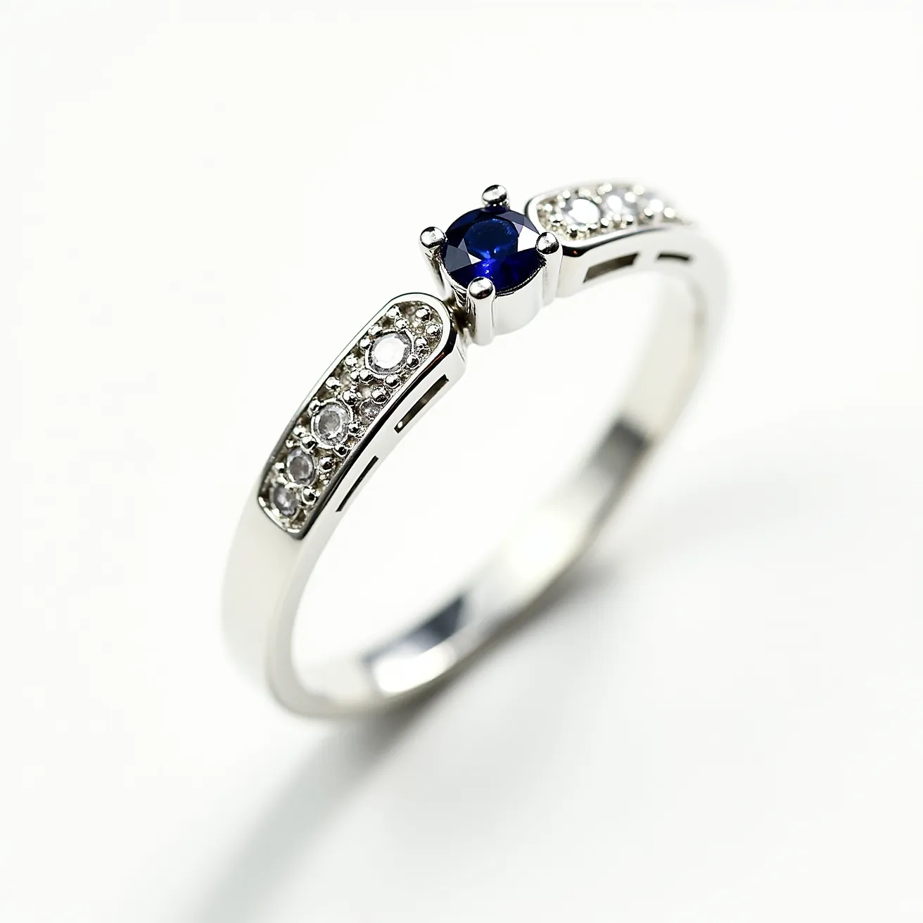 This pinky ring features a sleek design crafted from what appears to be white gold or platinum. At its center, a round-cut blue sapphire is secured in a prong setting, creating a striking focal point. Flanking the sapphire are small round diamonds, each set into the band in a pave setting, adding a touch of elegance and sparkle. The craftsmanship of the ring showcases a balance between the vibrant blue of the sapphire and the shimmer of the diamonds, making it both stylish and sophisticated.