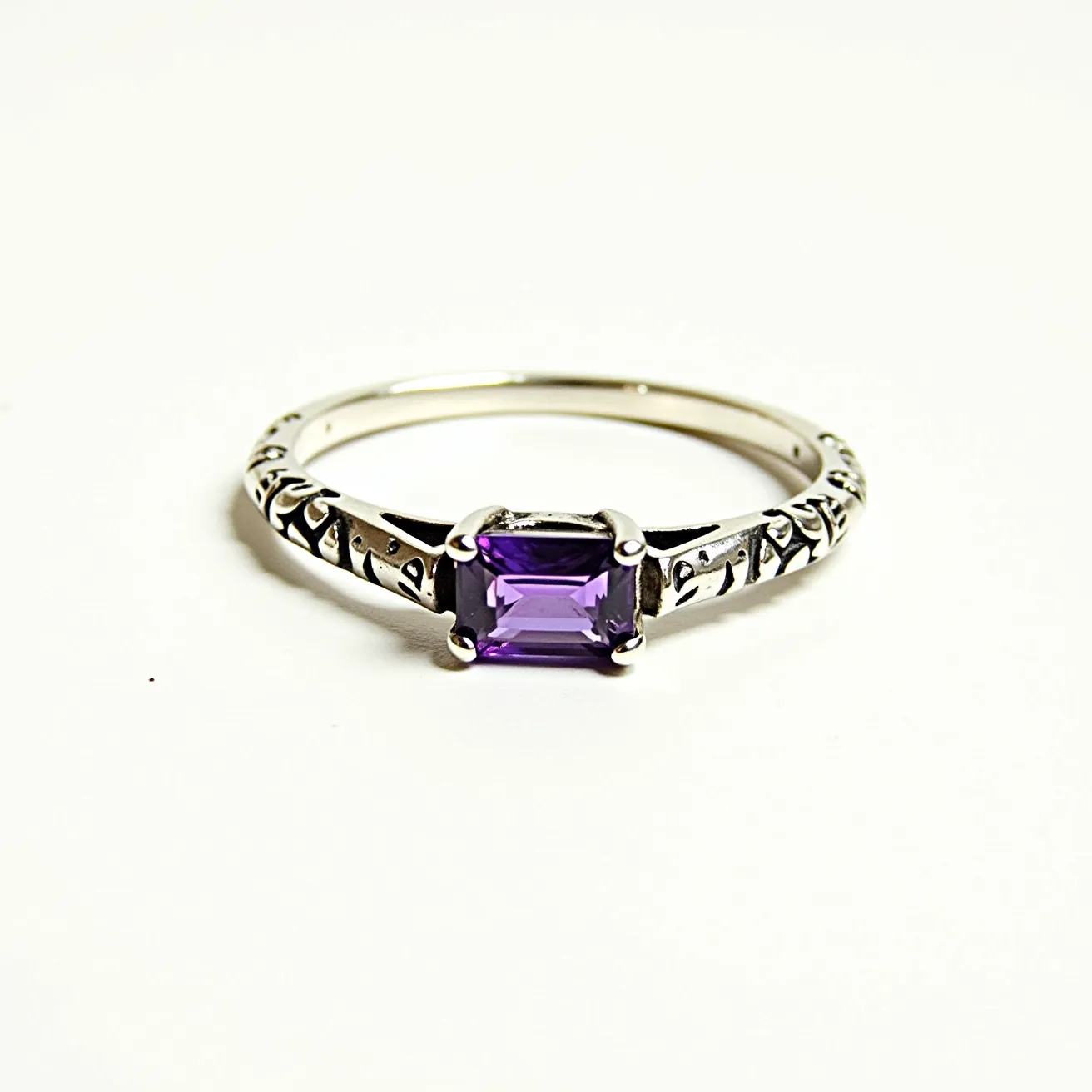 This pinky ring features a prominent rectangular-cut purple gemstone, likely an amethyst, securely held in a classic four-prong setting. The band appears to be crafted from a silver-hued metal, possibly sterling silver, and is adorned with intricate engraved designs that enhance its elegance. The prongs are designed for both security and style, ensuring the gemstone remains firmly in place. The overall craftsmanship suggests a focus on both aesthetic appeal and durability, making this ring a stylish and enduring adornment.