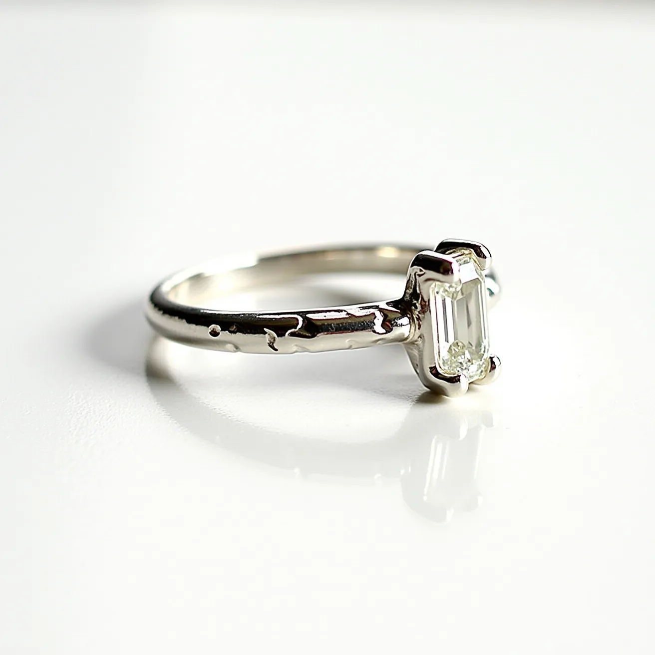 This pinky ring features a delicate design crafted from a silver-toned metal, which gives it a polished and refined appearance. It prominently showcases an emerald-cut gemstone set in a prong setting, allowing for a secure hold while displaying the stone's clarity and facets. The stone's rectangular shape adds an elegant aesthetic to the overall design. The band includes subtle textural details, adding a touch of uniqueness to the otherwise classic style of the ring.