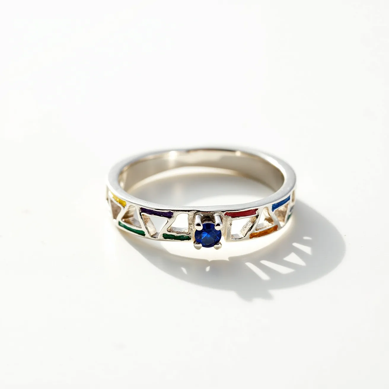 This rainbow ring features a sterling silver band adorned with a series of colorful enamel triangles set around its perimeter, showcasing red, orange, yellow, green, and blue hues. In the center, a round-cut blue gemstone is securely held in place by a four-prong setting, adding a focal point to the design. The silver band provides a sleek backdrop, enhancing the vibrancy of the colors and the brilliance of the central stone. 