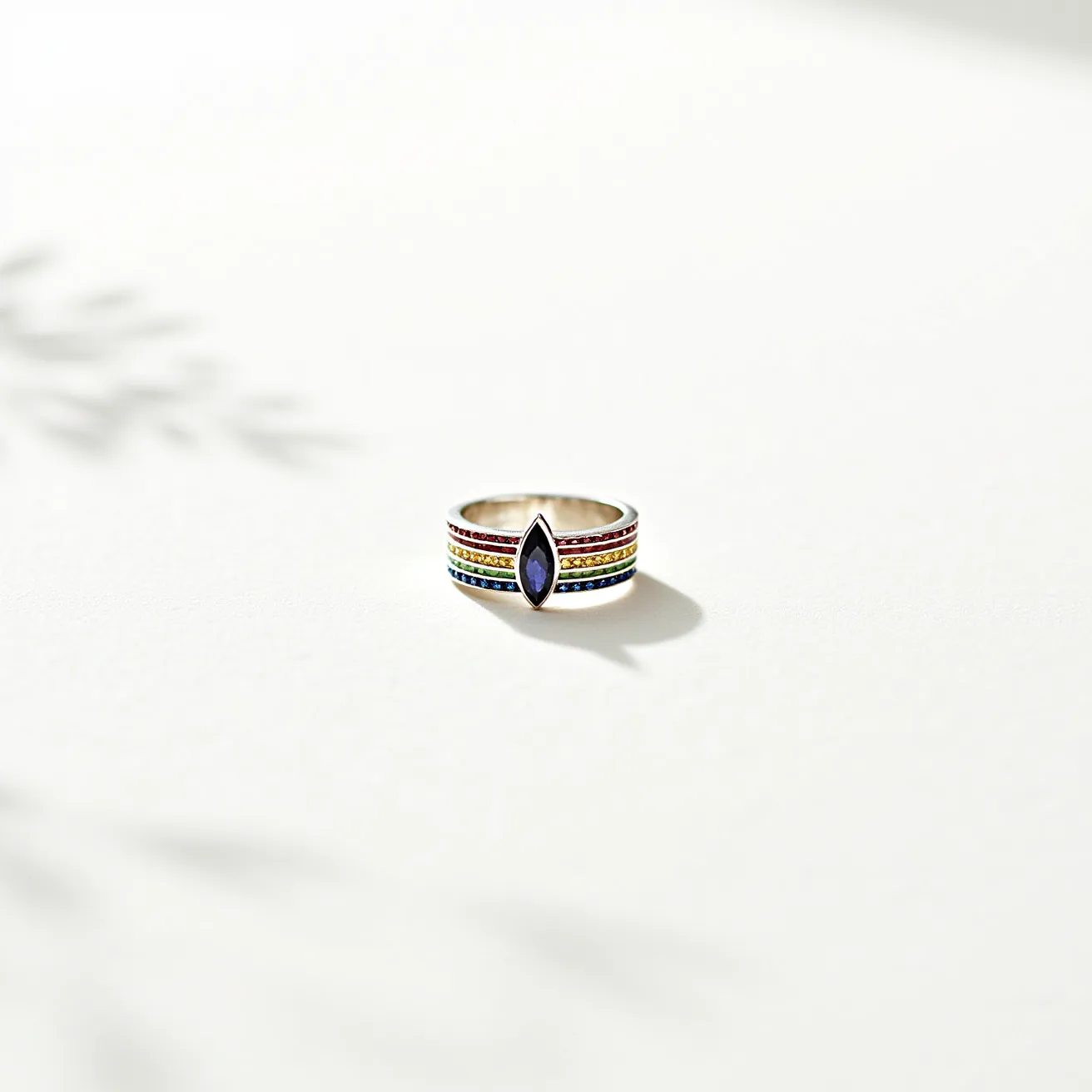 This rainbow ring features a stunning marquise-cut gemstone set vertically at its center, bordered by rows of small, multicolored gemstones that create a vibrant, rainbow effect across the band. The central stone is set in a bezel setting, providing a sleek and modern appearance. The band itself appears to be crafted from a shiny metal, possibly silver or white gold, enhancing the brightness of the multicolored stones. These smaller stones are round-cut and set in a pavé style to ensure a continuous and uninterrupted flow of color around the band, adding to the ring's elegant appeal.