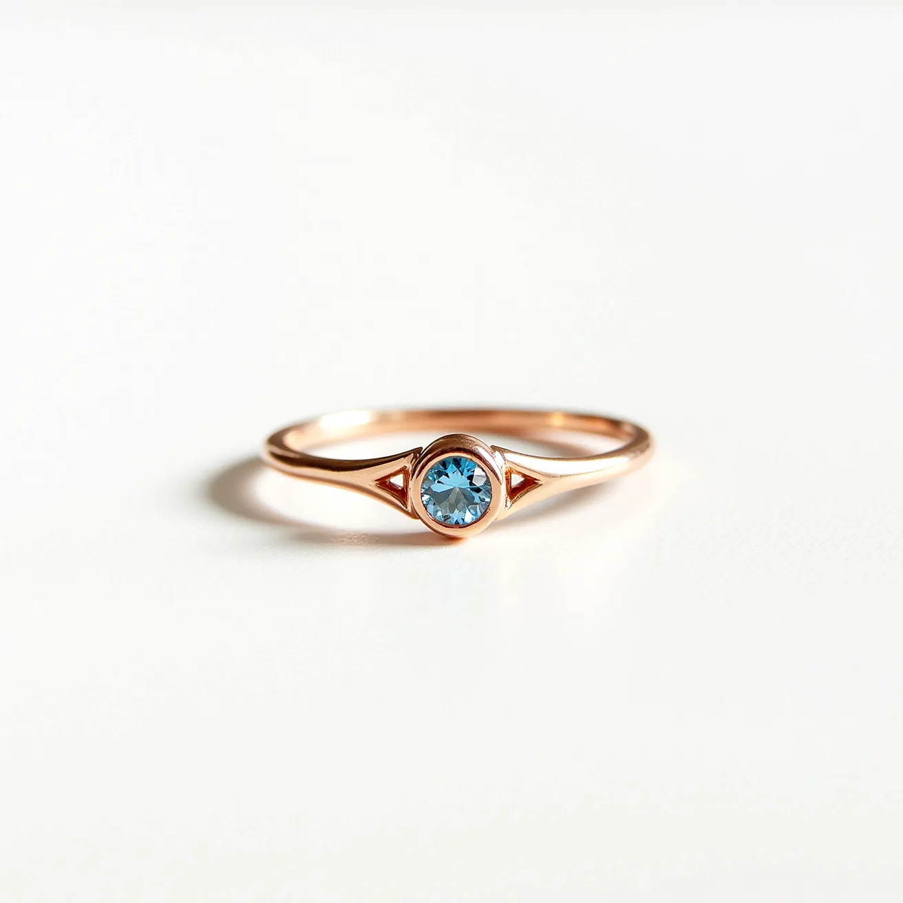 This Rapunzel ring features a delicate rose gold band, elegantly crafted to highlight its centerpiece, a round-cut blue gemstone. The gem is securely set in a bezel setting, which encircles it and provides a smooth, continuous line that enhances its vibrant color. Accent triangles flanking the central stone add a touch of sophistication, while the ring's simplicity and elegance make it a timeless piece suitable for various occasions.