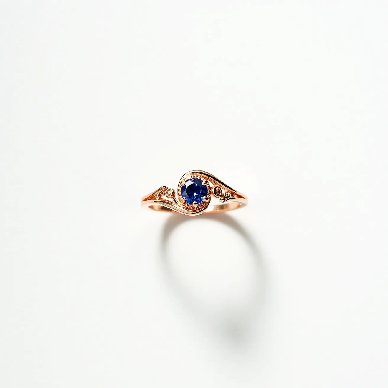 This Rapunzel ring features a central blue sapphire gemstone, cut in a round shape, and elegantly set in a prong setting that enhances its brilliance. The band is crafted from a rosy gold material that adds warmth and sophistication to the piece. Delicate scrollwork or engraved detailing adorns the band on either side of the central stone, adding an intricate, whimsical element to the overall design. The thoughtful combination of the vibrant sapphire and the pink-hued metal evokes a sense of fairy-tale charm and elegance.