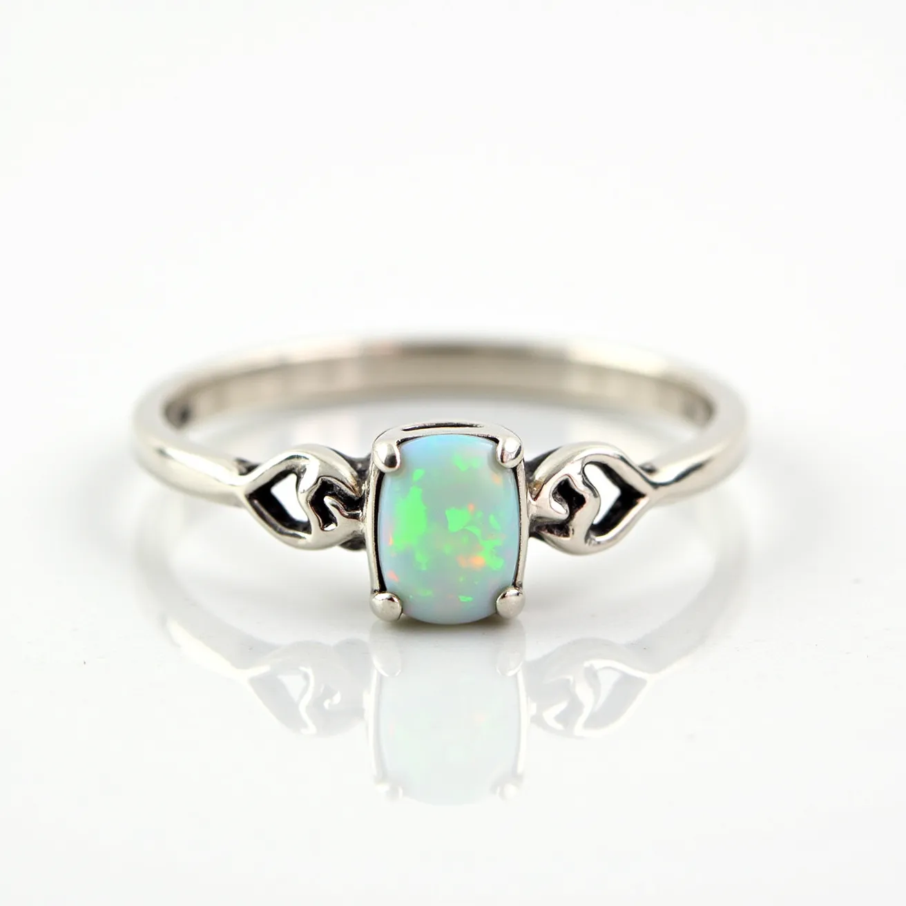 This ring band is crafted from a polished silver metal, featuring a delicate Celtic-inspired design on either side of a captivating centerpiece. At its heart lies an opal gemstone, characterized by its vibrant play of colors including greens and oranges. The opal is cut in a smooth, oval cabochon shape and is securely set with a classic four-prong setting that elegantly holds the gem in place. The combination of the detailed band design and the opal's iridescence gives the ring a distinct and enchanting appearance.