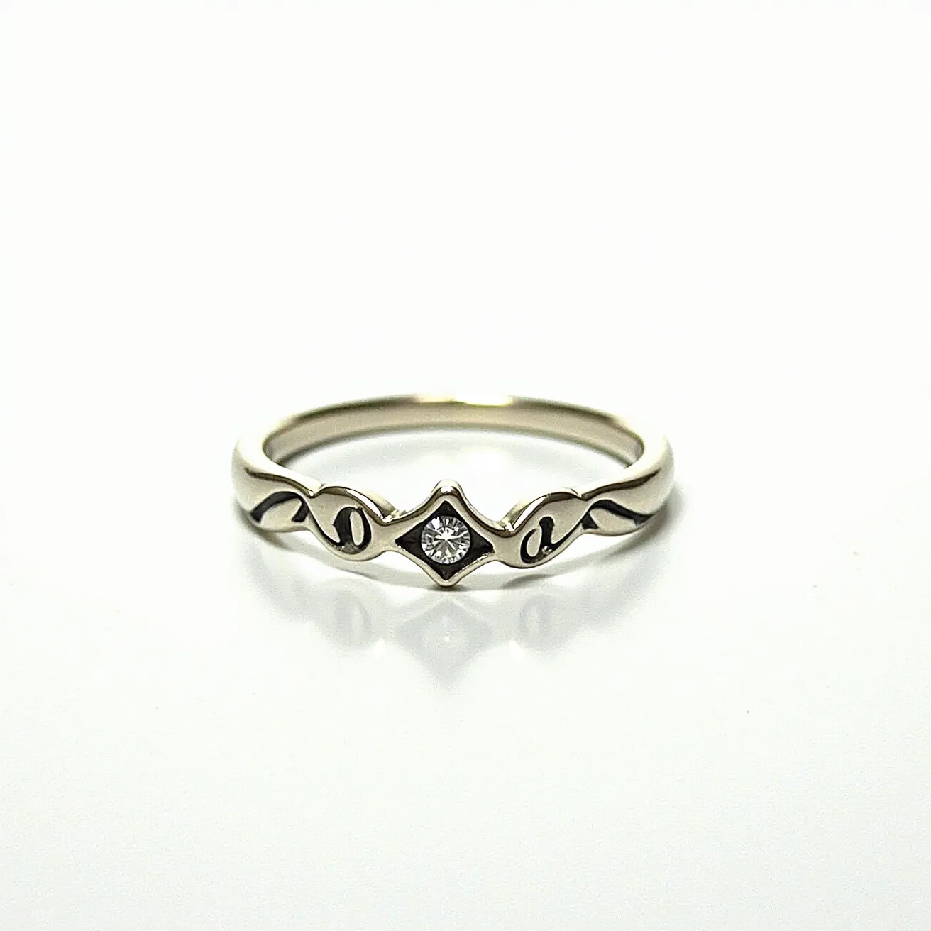 This ring band features a delicate and refined design crafted from a silver-toned metal. At its center, it showcases a single round-cut clear gemstone, likely a diamond or cubic zirconia, subtle in size and securely held in a bezel setting. The band is adorned with elegant, flowing engraved patterns on either side of the central stone, enhancing its sophisticated aesthetic. The ring does not include any additional clasps or attachments, maintaining a seamless and continuous design.