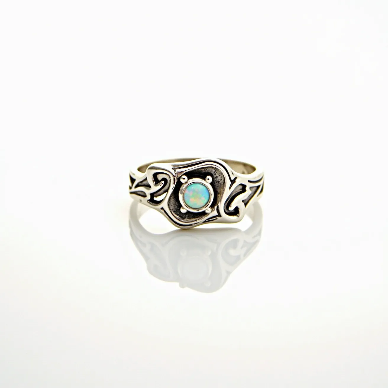 This ring band features a detailed design crafted from silver, highlighted by tribal-inspired engravings that give it a bold, artistic appearance. At the center, there is a round opal gemstone, which exhibits a captivating play of colors typical of opals. The opal is set in a bezel-style setting, securely framed within the metallic design. There are no additional clasps or attachments, as is typical for ring bands, emphasizing the smooth, continuous form that embraces the finger.