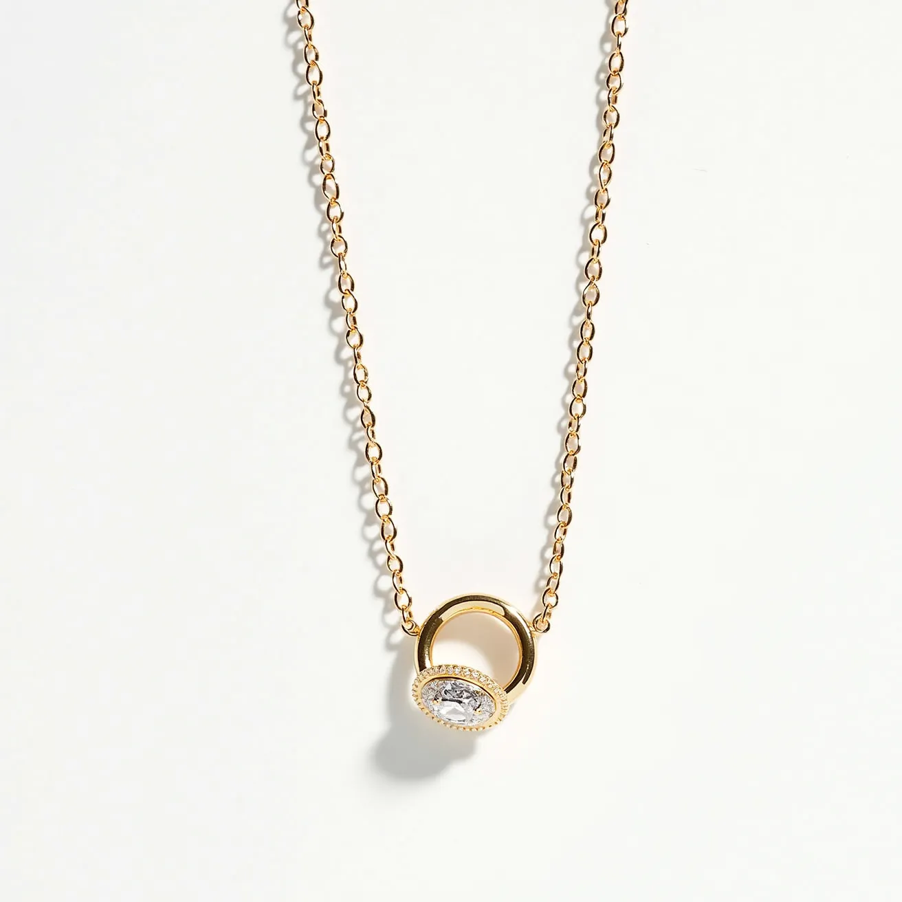 This ring necklace features a delicate gold chain that supports a gold ring pendant. Encrusted with small, clear gemstones around its surface, the central piece showcases an oval-cut stone securely set within the circular frame. The shimmering stones are prominently displayed, giving the piece an elegant and refined appearance. The attachment of the ring to the chain appears seamless, enhancing the overall sleek and cohesive design of the necklace.