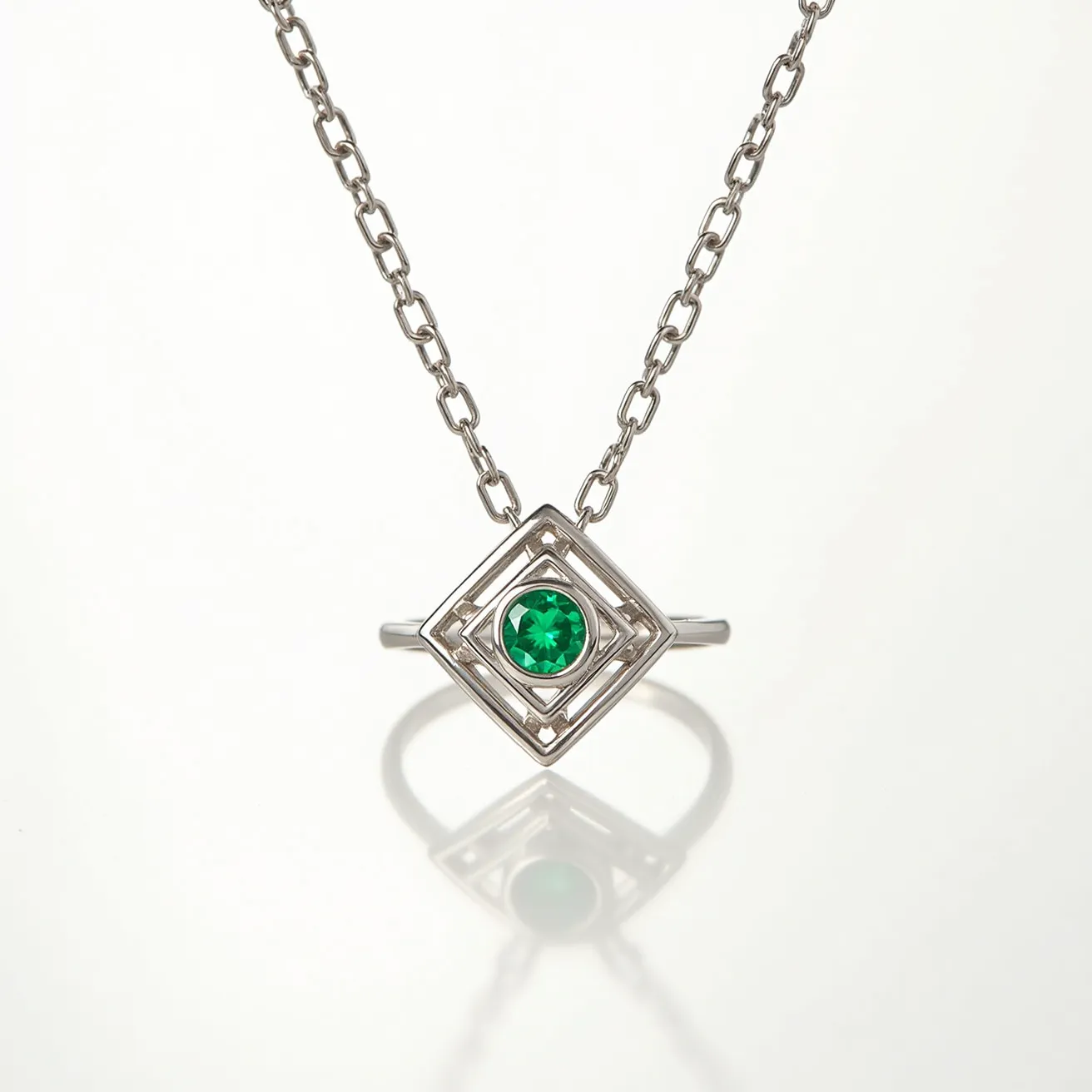 This ring necklace features a striking design where a circular band is integrated with a square-shaped decorative element at its center. It is crafted from a metallic material with a polished silver appearance, which forms both the chain and the geometric motifs. At the center of the square sits a vibrant green gemstone, cut into a round shape and secured in a bezel-style setting, accentuating its color and brilliance. The chain is composed of small, uniform links that provide a balanced and elegant aesthetic. The necklace does not appear to have a visible clasp, likely integrating a discreet fastening solution within the chain or pendant design.