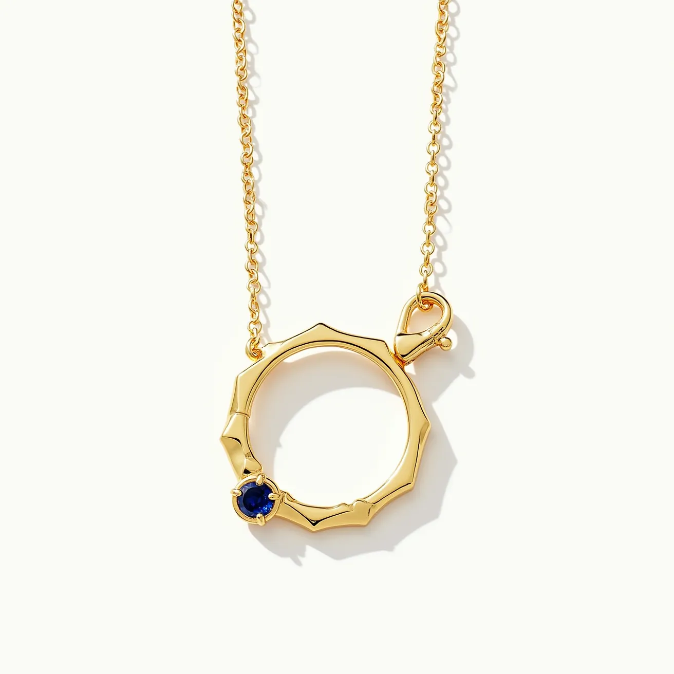 This ring necklace features a gold-toned metal with a polished, angular design, creating a bold and modern aesthetic. The centerpiece of the necklace is a round, blue gemstone set into the ring, likely a sapphire, enhancing the luxurious feel with its deep hue. The stone is held securely in a prong setting, which allows light to pass through and highlight its color. The necklace is equipped with a lobster clasp attached to the ring, providing a secure and user-friendly fastening mechanism. The chain is a delicate, matching gold-toned link design, complementing the overall elegant appearance of the piece.