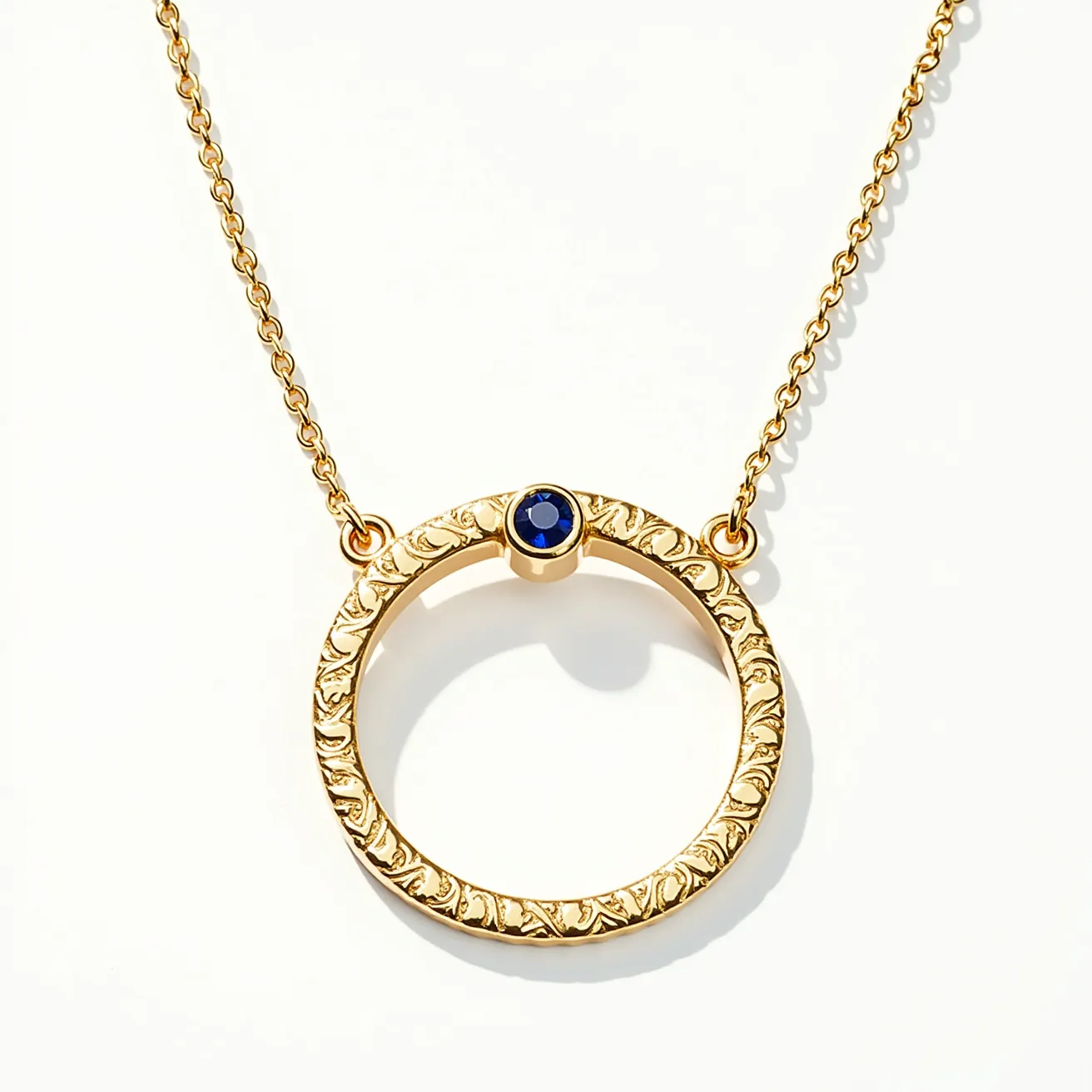 This ring necklace features a gold-tone circular pendant with intricate embossed detailing, providing a textured and elegant appearance. Set into the top of the circle is a deep blue gemstone, likely a sapphire, with a round cut that adds a touch of sophistication. The stone is securely held in place with a bezel setting, ensuring durability and style. The pendant is linked to the necklace chain through small gold loops on either side, seamlessly integrating into the overall design. The chain itself appears to be a delicate link chain, which complements the pendant's refined look.