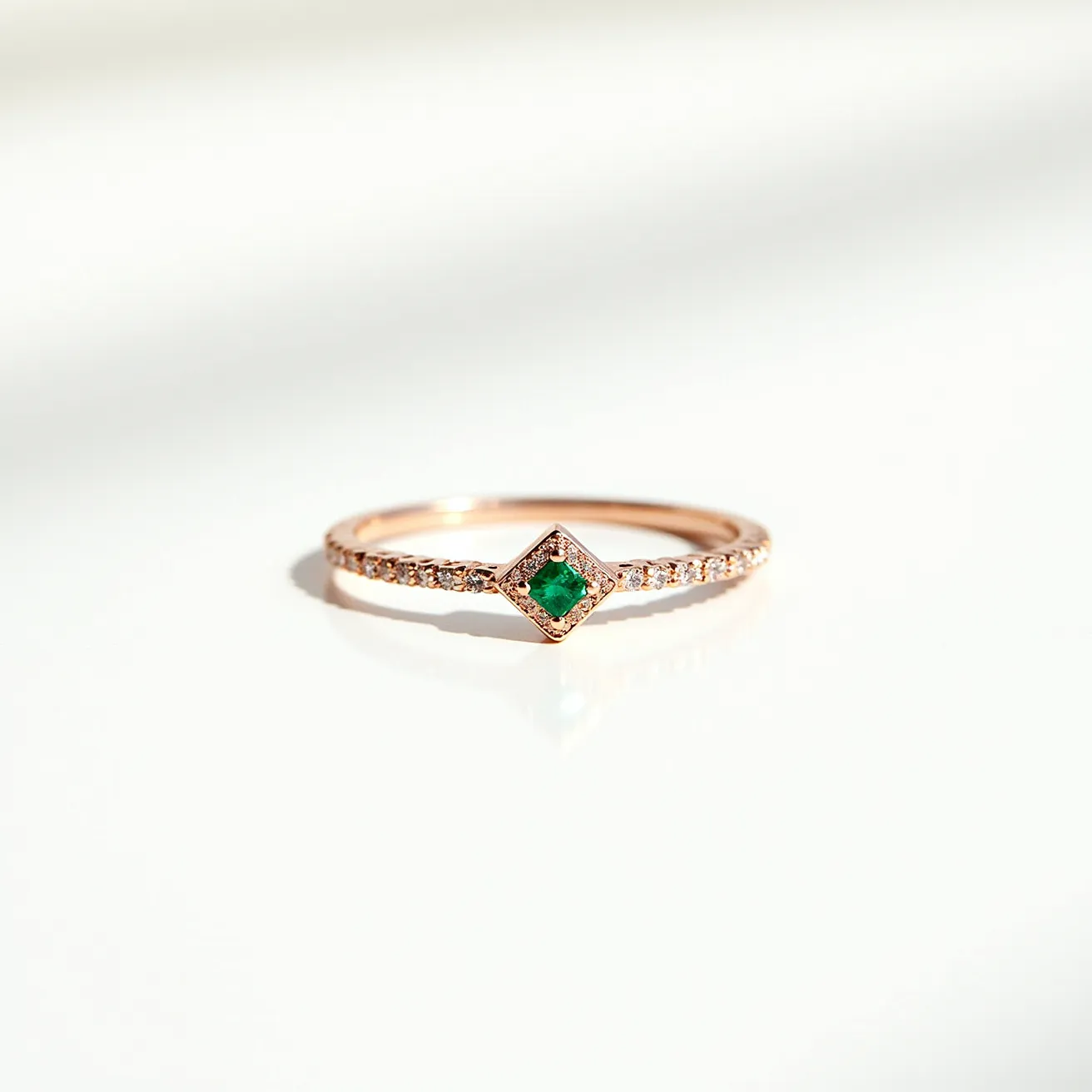 This rose gold ring features a delicate design, characterized by a central green gemstone set in a distinctive diamond shape. The stone appears to be an emerald with a round cut, adding a vibrant touch of color. Flanked by a row of small, round, clear stones likely set as pavé on the band, these complementary stones enhance the ring's overall sparkle and elegance. The minimalist band is crafted from rose gold, providing a warm, classical backdrop that accentuates the gems' brilliance. The design omits any additional clasps or attachments, focusing instead on the seamless integration of these elements to create a harmonious and elegant piece.