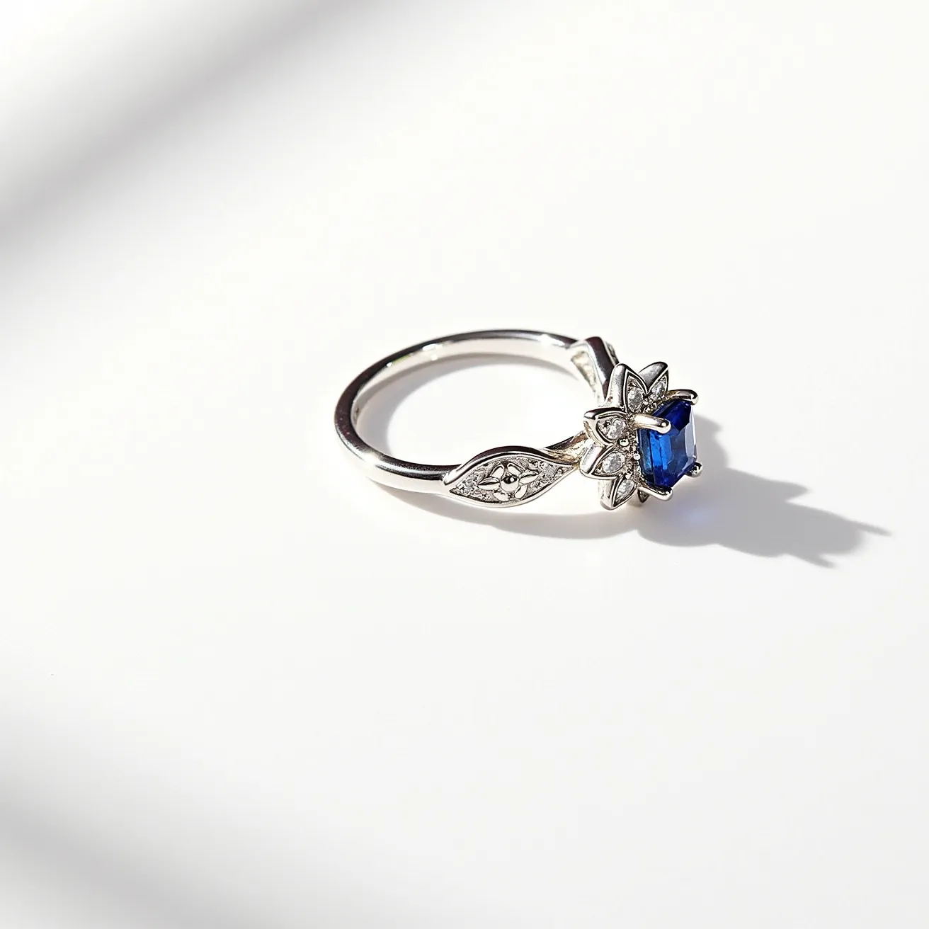 This rose ring features a delicate design crafted from what appears to be silver or white gold, showcasing a central blue sapphire gemstone. The sapphire is an emerald cut, nestled in a subtle floral setting that enhances its vibrant hue. Surrounding the central stone are small, round accent diamonds set in a pavé style, adding a touch of sparkle. The band is elegantly simple with a decorative element resembling leaves, further enhancing the floral motif of the ring. The design is seamless, with no visible clasps or attachments, allowing for a smooth and comfortable fit.