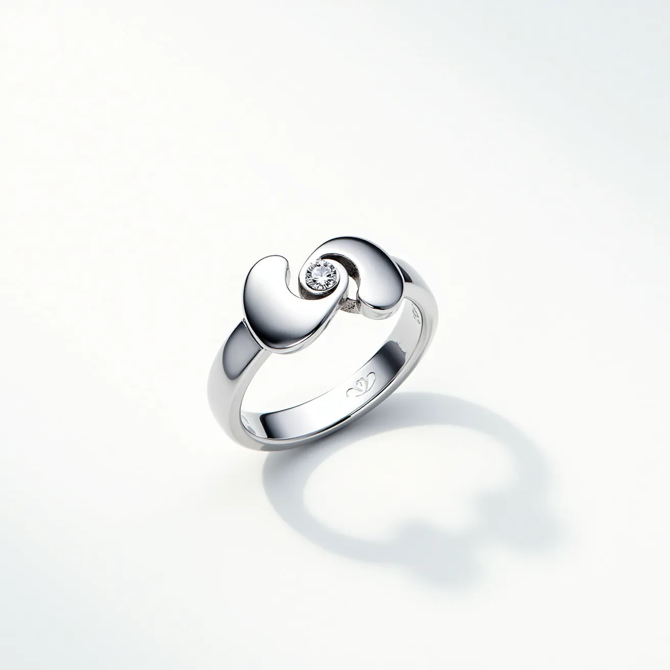 This rose ring features a sleek metal band that appears to be crafted from polished silver or white gold, providing a modern and elegant look. At the center, it showcases a single round-cut diamond set within the band, secured by bezel settings. The design incorporates two gracefully interlocking curves that accentuate the central diamond, enhancing the modern aesthetic. The interior of the band is smooth, ensuring comfortable wear, and is likely to have a hallmark or brand engraving. The ring’s composition and design demonstrate a refined combination of simplicity and sophistication.