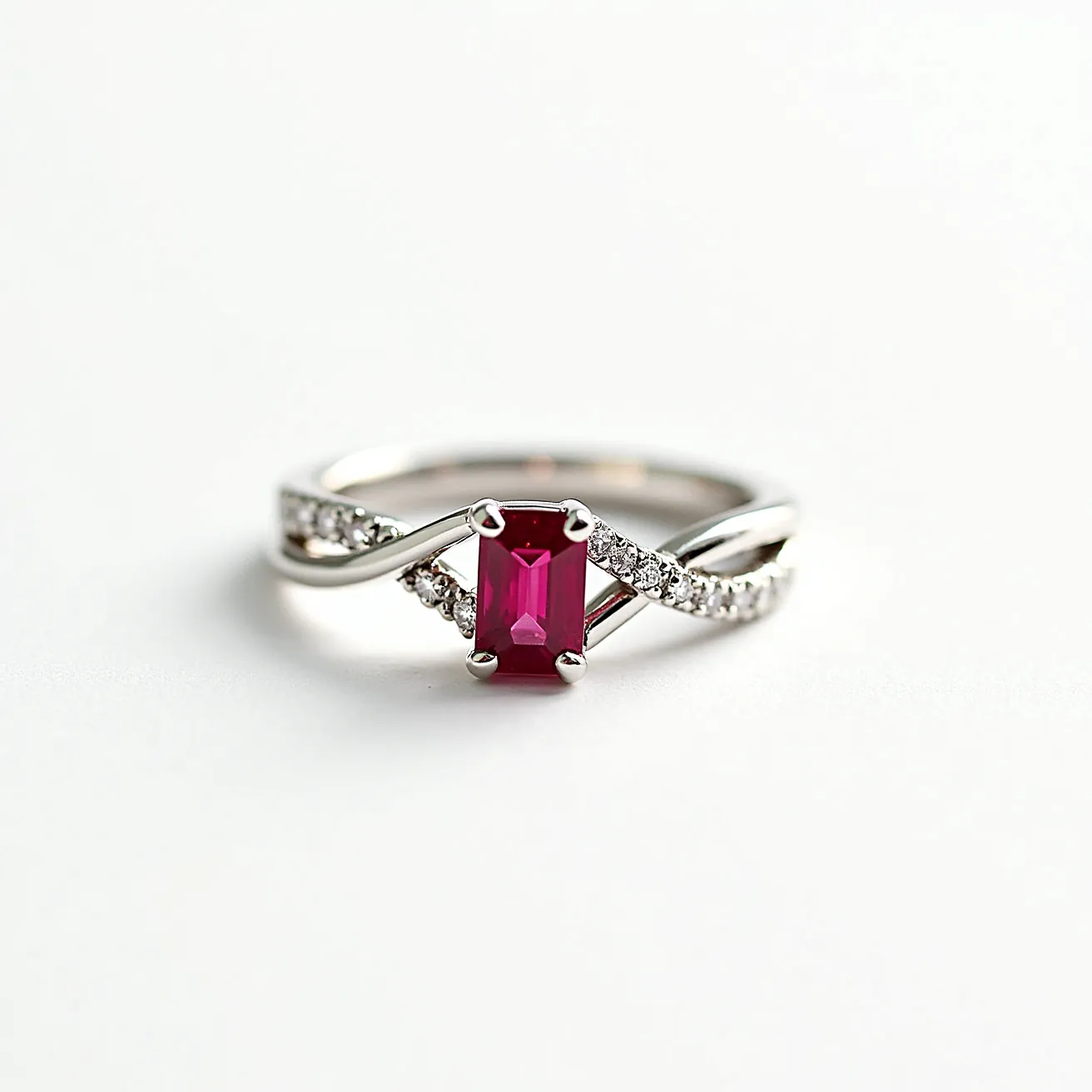 This ruby engagement ring features a striking emerald-cut ruby as its centerpiece, set in a classic prong setting that highlights its vibrant red hue. The band is crafted from polished white gold, showcasing an elegant intertwined design that adds a touch of modern sophistication. Along the band, accentuating the central ruby, are small round brilliant-cut diamonds, pave-set to enhance the overall sparkle and elegance of the ring. The setting and band combine timeless elegance with a contemporary twist, making this piece both unique and captivating.