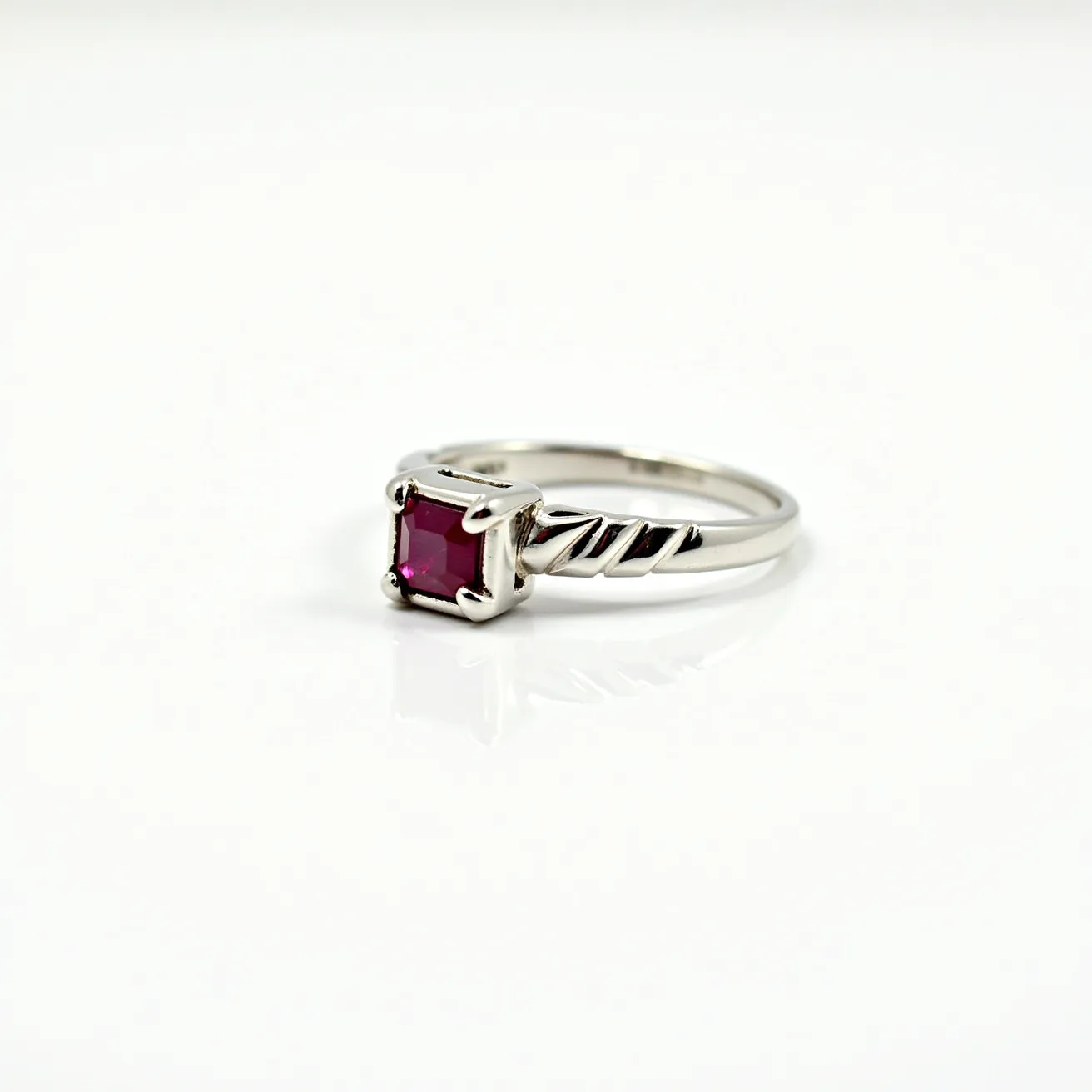 This ruby ring features a square-cut ruby, held securely by four prongs in a classic setting. The band appears to be crafted from silver or white gold, providing a sleek and elegant look. The band also displays intricate twisted detailing, adding a touch of sophistication and unique style to the piece. The setting allows the ruby to be the central focus, showcasing its vibrant red hue. The ring does not feature any additional attachments or clasps, emphasizing its minimalist and refined design.