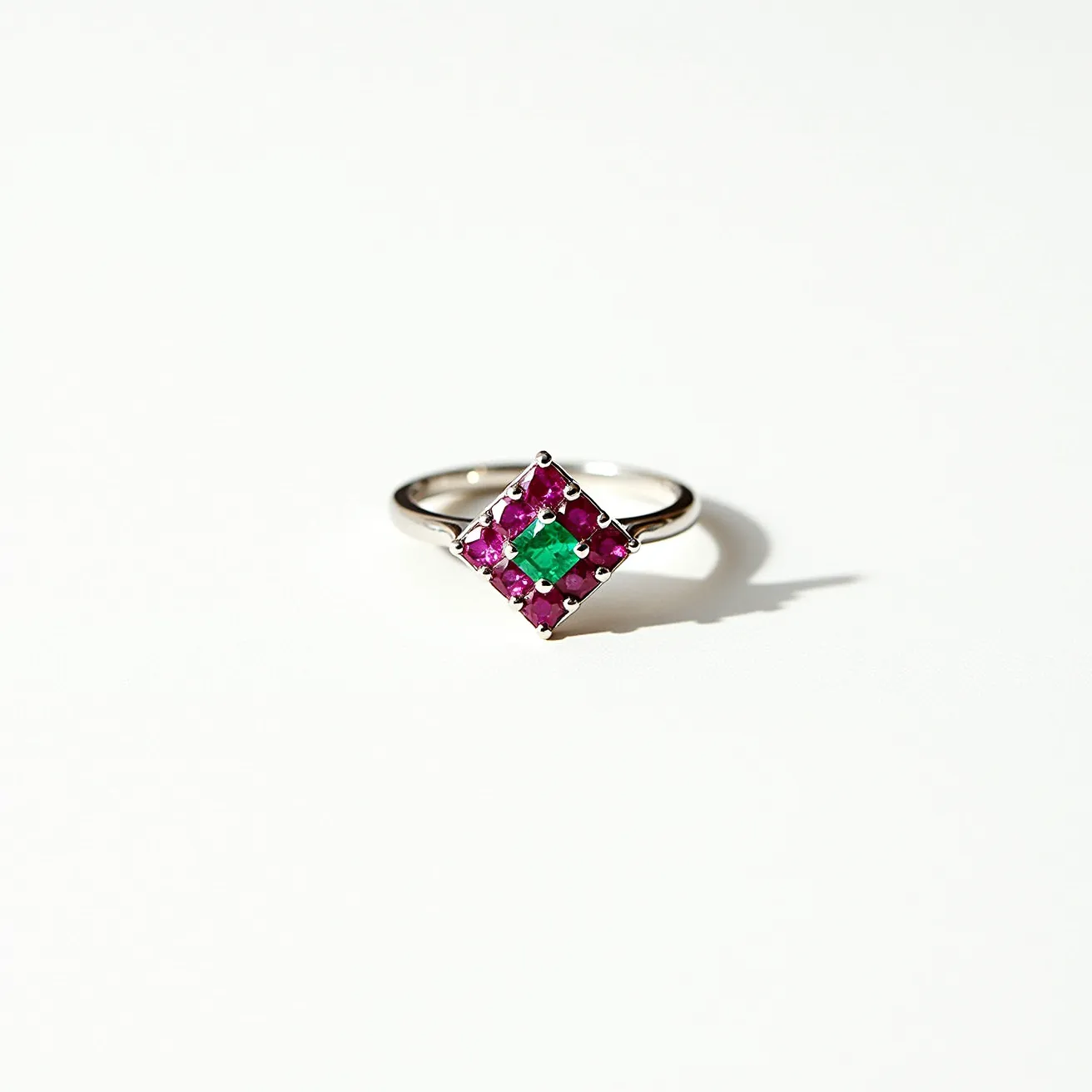 This ruby ring features a vibrant green gemstone set in the center, surrounded by vividly colorful pink gemstones arranged in a square pattern, creating a striking contrast. The stones are mounted on a sleek, silvery band, which could be made from platinum or white gold, known for enhancing the brilliance of gemstones. The central stone is likely an emerald, cut in a square shape, with rubies around it in a similar cut, offering a classic and elegant appearance. The setting appears to be a prong setting, securely holding each stone while allowing maximum light to showcase their brilliance.