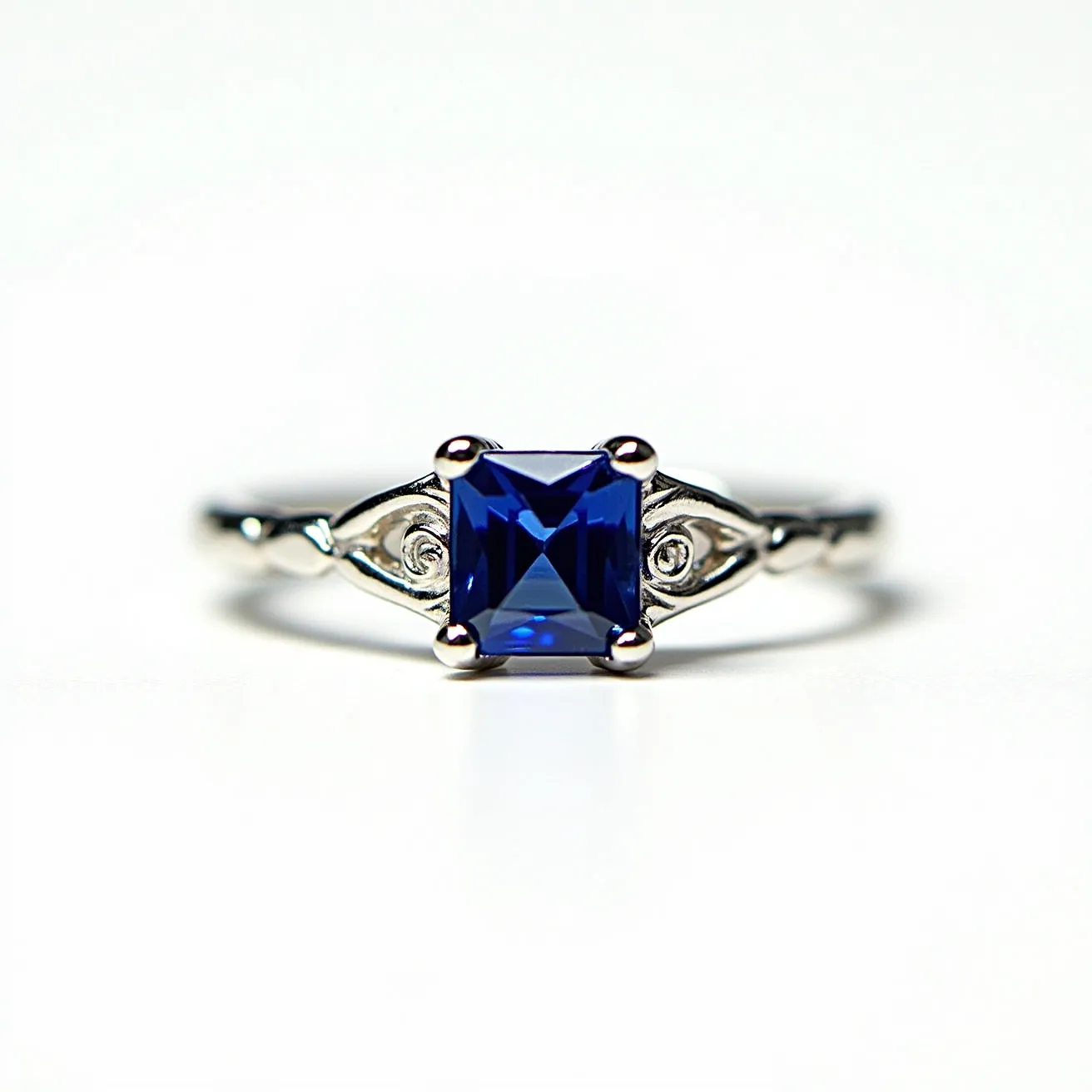 This sapphire ring features a striking square-cut blue sapphire as its centerpiece, held securely in a prong setting that accentuates the gem’s brilliance. The band appears to be crafted from polished white metal, possibly white gold or platinum, enhancing the sapphire's deep blue hue. Elegant detailing flanks the central stone, with small, circular accents adding a touch of sophistication on each side. The design is both modern and timeless, making it a versatile piece of jewelry.