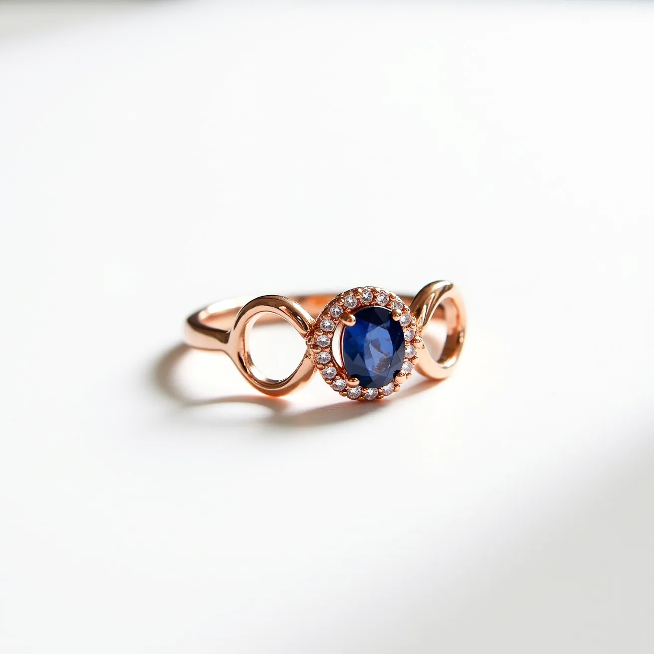 This sapphire ring features a striking oval-cut blue sapphire at its center, encircled by a halo of small, round brilliant-cut diamonds. The stones are securely set in a rose gold band that incorporates an elegant open loop design on either side of the central setting, adding an artistic touch to the overall appearance. The combination of the deep blue of the sapphire and the sparkling diamonds enhances the visual appeal, making it a distinctive and elegant piece of jewelry.