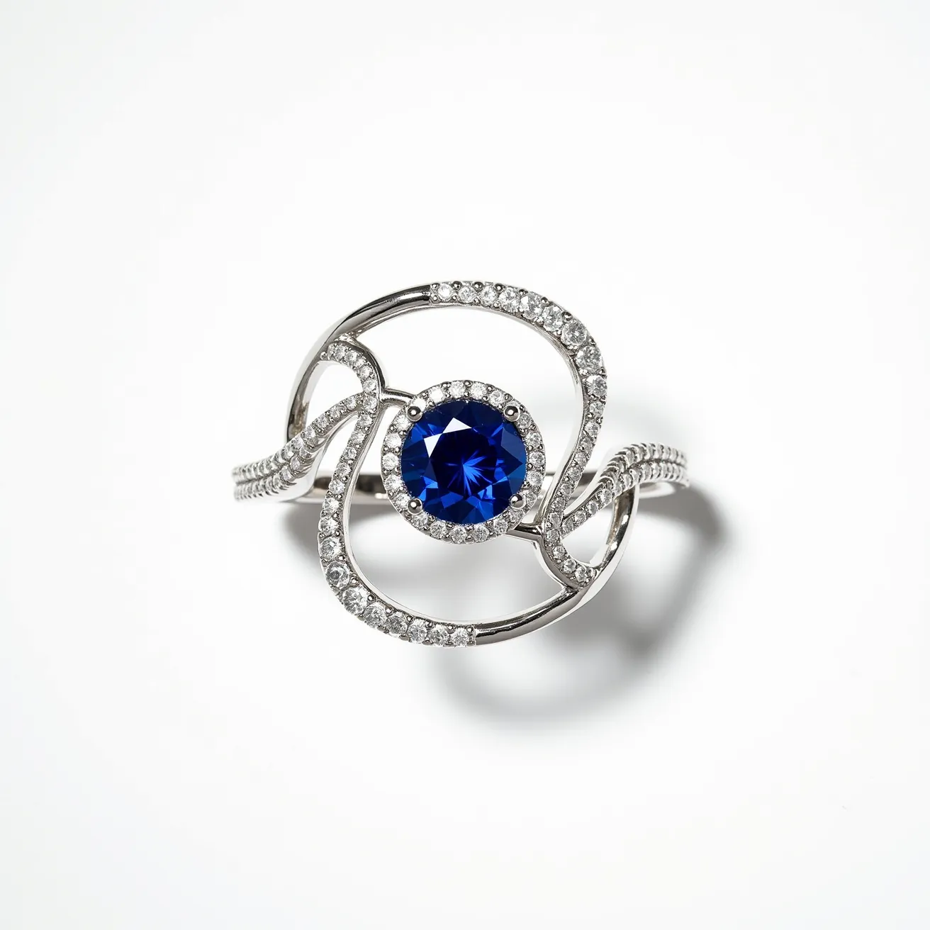 This sapphire ring features a stunning deep blue, round-cut sapphire set at its center. The sapphire is surrounded by a halo of smaller, brilliant-cut diamonds, enhancing its vibrant color and brilliance. The intricate band appears to be crafted from white gold or platinum, elegantly looping around the sapphire and encrusted with additional diamonds, adding to the overall sparkle and design of the piece. The setting demonstrates a creative, artistic flow that integrates the stones seamlessly into the metalwork, showcasing the craftsmanship and elegance of the ring design.