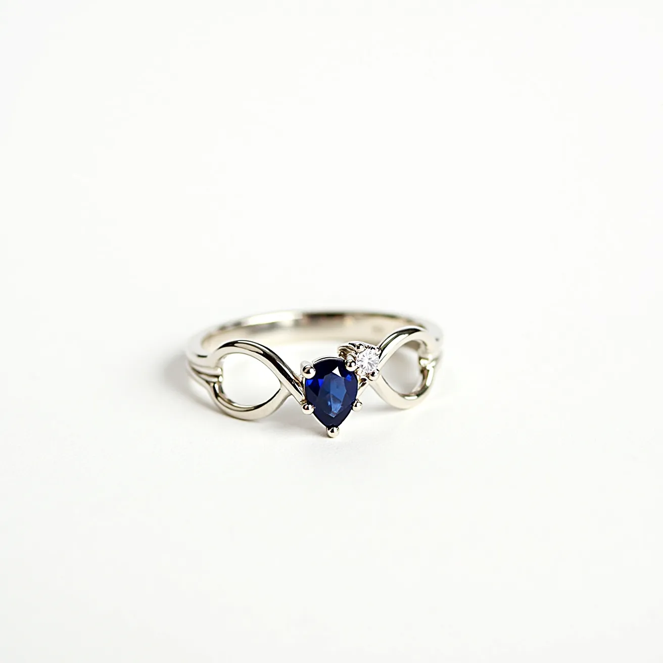 This sapphire ring features a striking, heart-shaped blue sapphire as its central gem, intricately set within a fluid, infinity-shaped band. Accompanying the sapphire is a small, brilliant-cut diamond positioned to the side, adding an elegant sparkle to the overall design. The ring band appears to be crafted from a lustrous white metal, likely white gold or platinum, complementing the gemstones' brilliance and enhancing their vibrant hues. This exquisite piece embodies a harmonious blend of classic elegance and modern style, marked by its graceful lines and thoughtful gemstone arrangement.