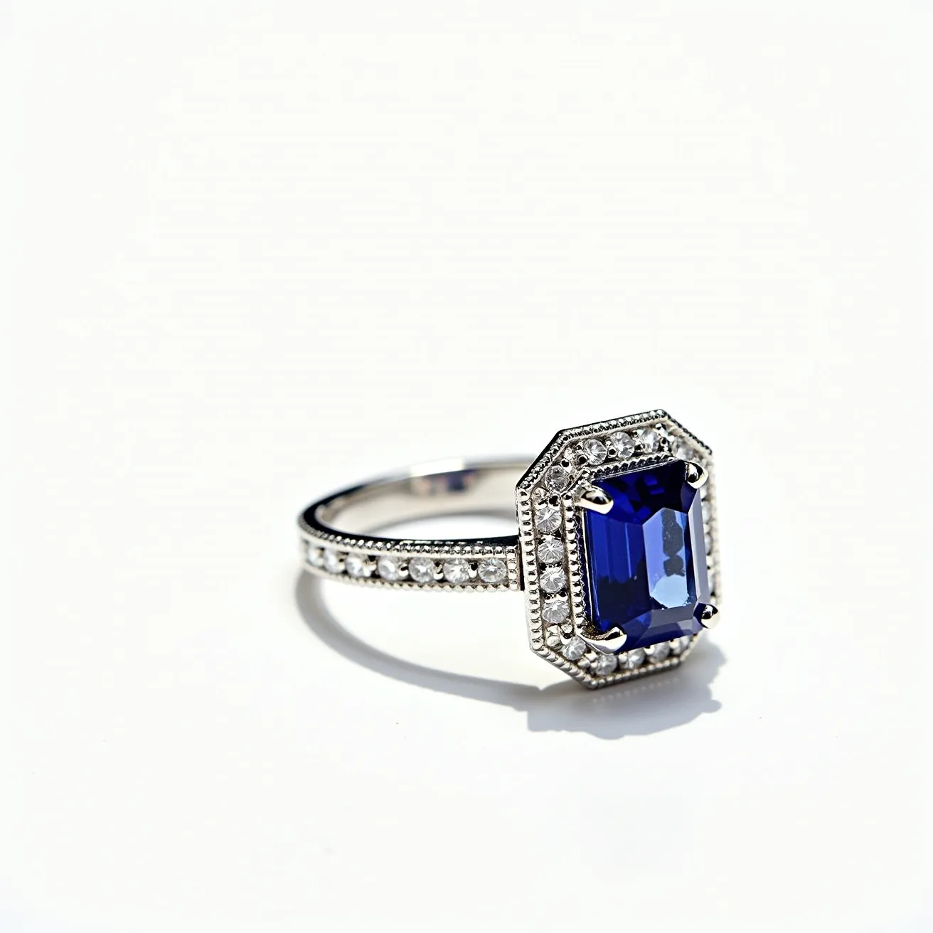 This sapphire ring features a stunning deep blue sapphire as the centerpiece, cut in an emerald shape and securely held in place by a four-prong setting. Encircling the sapphire is a halo of small, round-cut diamonds, which add a brilliant sparkle to the piece. The band itself is crafted from a polished metal, likely platinum or white gold, and is adorned with additional small diamonds, adding to the ring's elegance and luxurious feel. The intricate detailing on the band enhances its visual appeal, creating a sophisticated and timeless design.