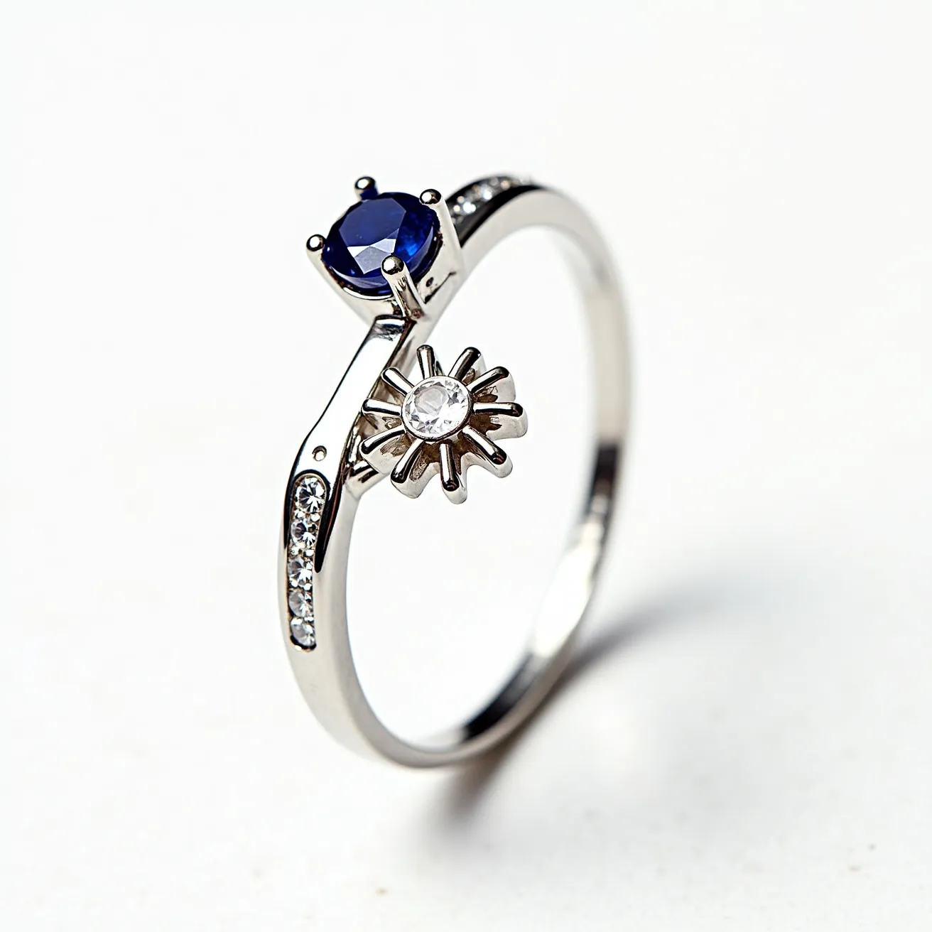 This sapphire ring features an elegant design, prominently showcasing a deep blue, round-cut sapphire as its centerpiece, held securely in place by a classic prong setting. Adjacent to the sapphire is a decorative floral element, which includes a clear, round-cut gem at its center, adding a whimsical touch to the ring's design. The band, likely made of polished white gold or platinum, is adorned with a series of small, clear stones set into its surface, enhancing the overall sparkle and sophistication of the piece. The ring exhibits meticulous craftsmanship, with each element seamlessly integrated to create an enchanting and stylish accessory.