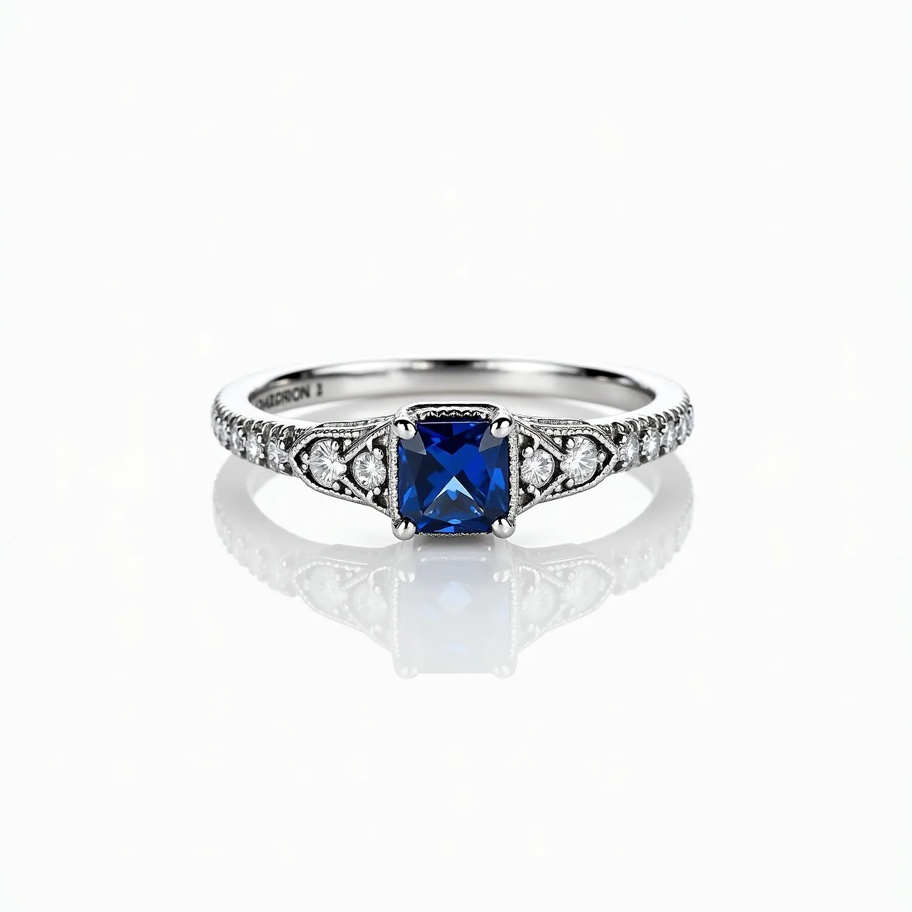 This sapphire ring features a striking square-cut blue sapphire as its centerpiece, set in a prong setting that secures the gemstone while allowing maximum light reflection. The band is crafted from a lustrous white metal, likely platinum or white gold, and showcases intricate detailing with a series of small, round-cut diamonds embedded along the shoulders, adding a touch of sparkle and elegance. The ornamental design on either side of the sapphire enhances the ring's sophistication, while the overall setting emphasizes the vibrant blue of the sapphire.