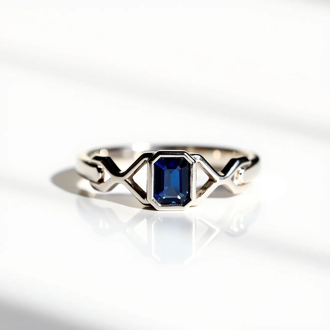 This sapphire ring features a striking blue emerald-cut sapphire set at its center, creating a focal point of elegance. The sapphire is securely held in a bezel setting, which enhances its geometric shape and provides a modern yet classic appeal. The band of the ring is crafted from a polished metal, likely white gold or platinum, which complements the deep blue of the gemstone beautifully. The design of the band is unique, with geometric patterns adding a distinctive style to the piece. This combination of materials and design elements gives the ring a sophisticated and timeless look.