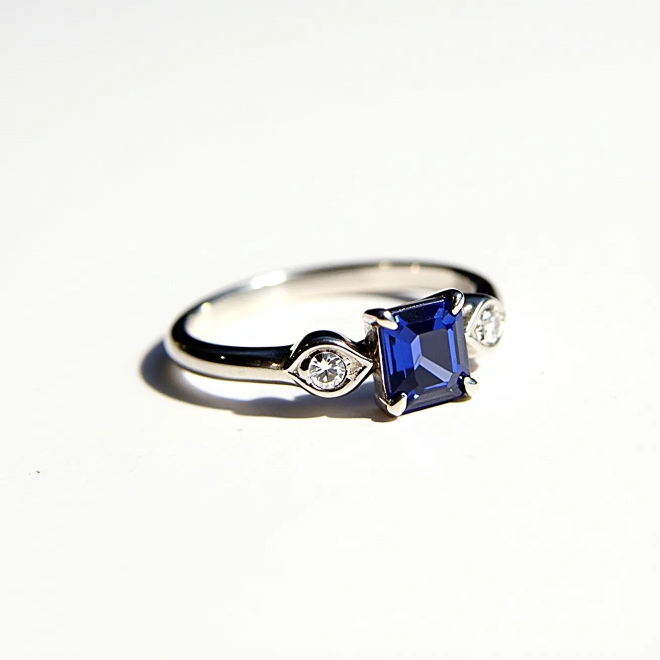 This sapphire ring is elegantly designed with a square-cut blue sapphire as the centerpiece, set in a prong setting that showcases its vibrant color and clarity. Flanking the central stone are two round brilliant-cut diamonds, adding a touch of sparkle and balancing the design. The band is crafted from a lustrous metal, likely platinum or white gold, which enhances the brilliance of the gems and provides a modern, sophisticated look. The overall composition of the ring is sleek and refined, with a timeless appeal suited for various occasions.