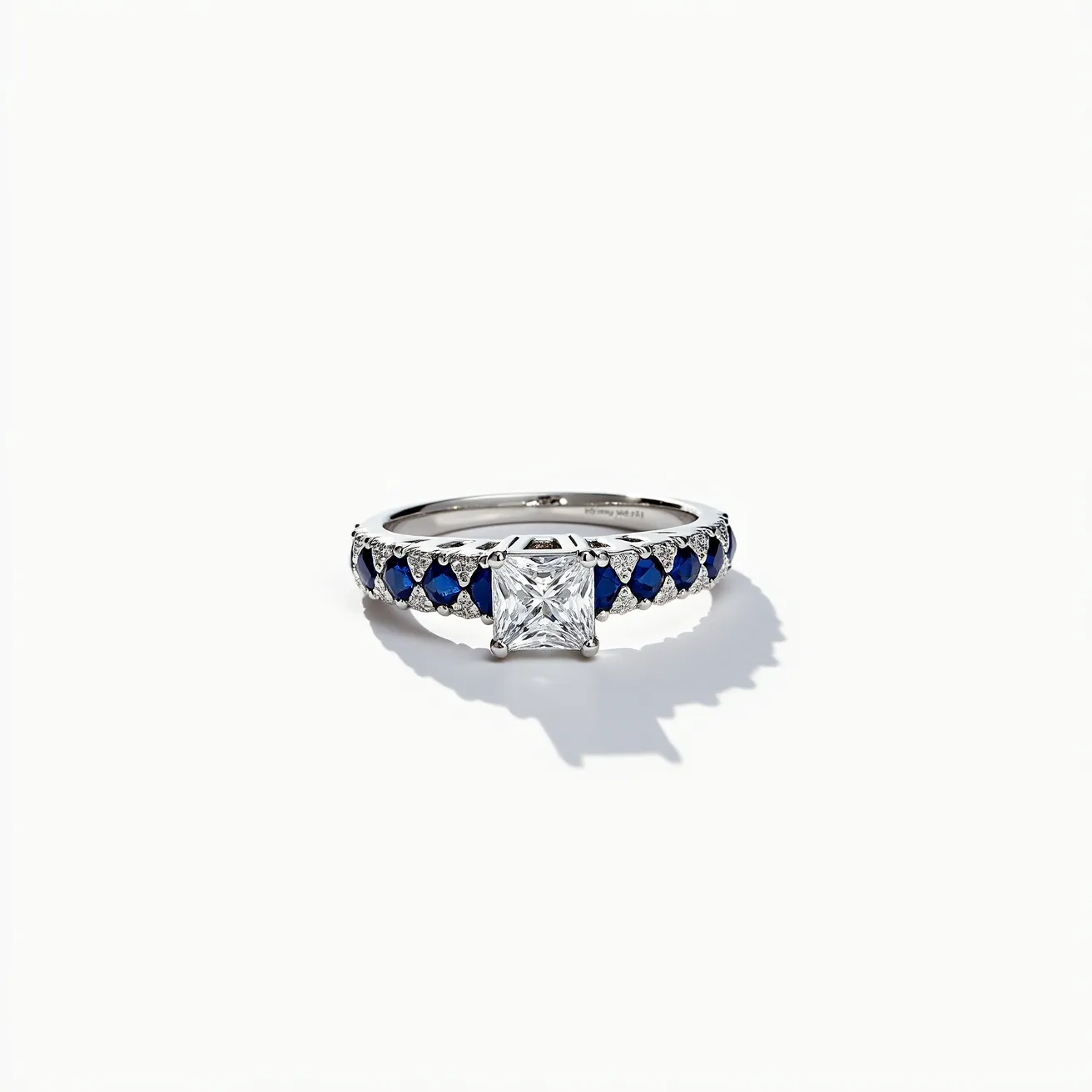 This sapphire ring features a central princess-cut diamond set in a classic prong setting, flanked by a series of deep blue sapphires along the band. The sapphires are arranged in a pattern that alternates with smaller round diamonds, adding a touch of sparkle and contrast. The metal of the band appears to be a reflective white material, likely platinum or white gold, providing a sleek and elegant finish. The ring is designed as a continuous loop without any visible clasp or attachment, emphasizing the seamless and luxurious style of the piece.