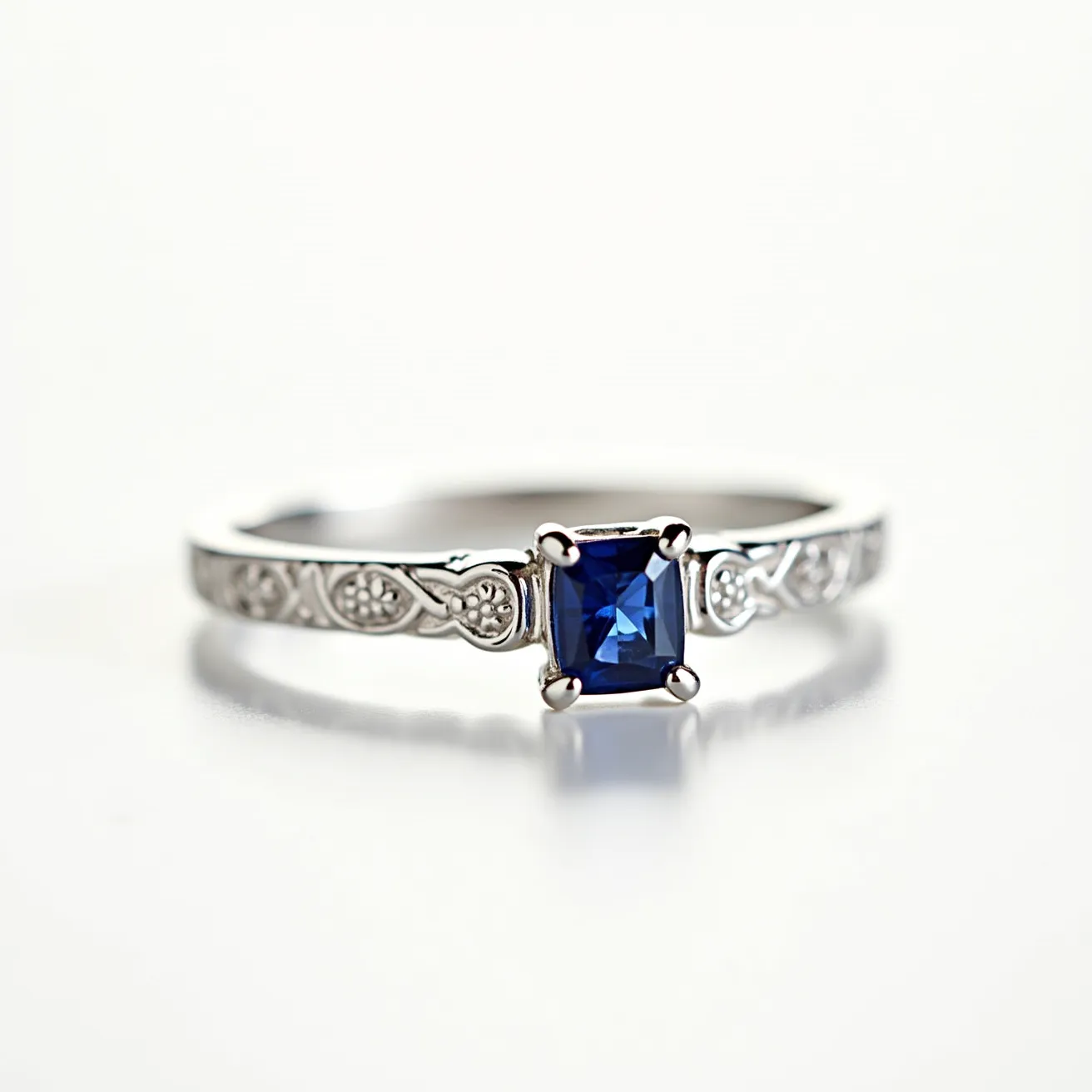 This sapphire ring features a striking square-cut blue sapphire as its centerpiece, securely held by a four-prong setting. The band appears to be made of a white metal, potentially platinum or white gold, and is adorned with intricate engraved patterns, adding a touch of elegance. The craftsmanship highlights the contrast between the deep blue sapphire and the shimmering band, creating a classic and sophisticated piece of jewelry.