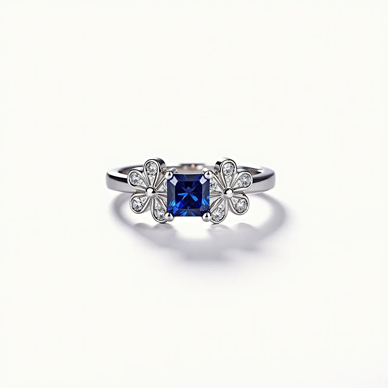 This sapphire ring features a striking square-cut sapphire as its centerpiece, nestled in a sophisticated prong setting. The band is likely crafted from a high-quality metal, possibly platinum or white gold, enhancing the ring's overall elegance. Flanking the central sapphire, there are intricate floral designs encrusted with smaller round-cut diamonds, which add a delicate brilliance and complement the deep blue of the sapphire. The combination of gemstones and design elements contributes to a luxurious and unique appearance, making the ring a true statement piece.