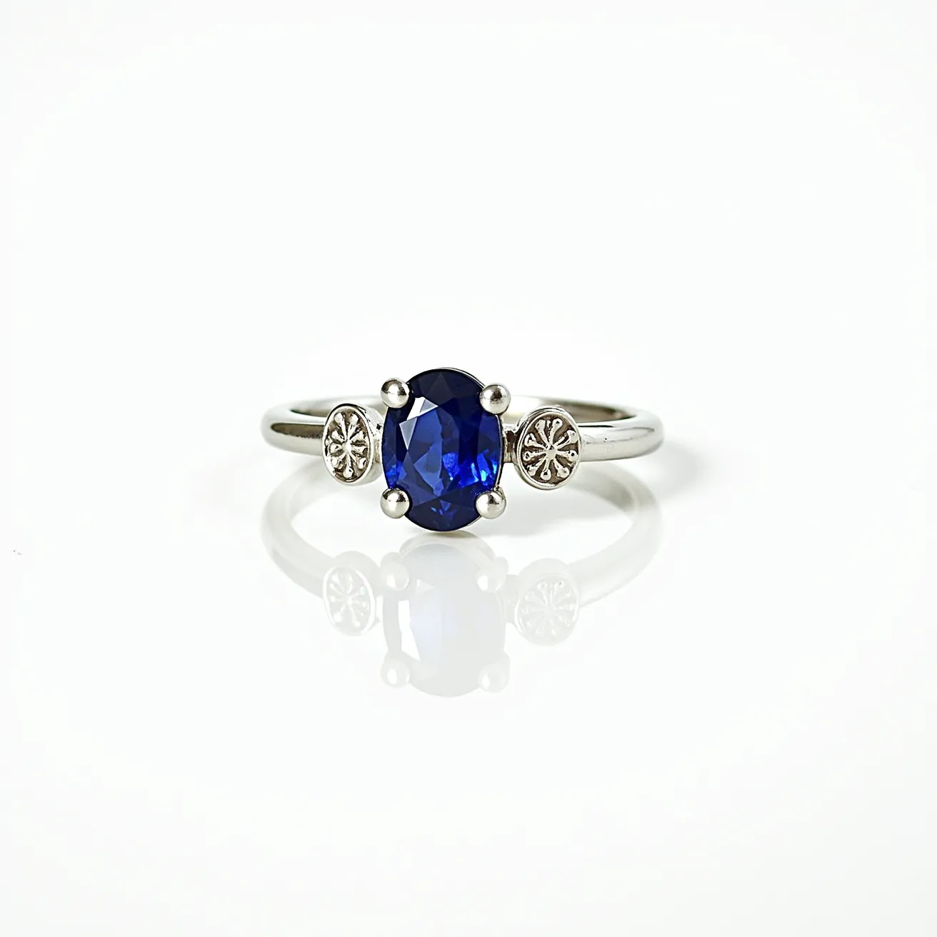 This sapphire ring features a vibrant oval-cut blue sapphire set in a sleek, polished metal band, likely crafted from a precious metal such as white gold or platinum. The sapphire is held securely in a four-prong setting, ensuring both elegance and stability. Flanking the central stone are two small, intricately designed discs, adding a touch of unique artistry to the overall design. The clean lines and refined details of the ring enhance its sophisticated and timeless appeal.