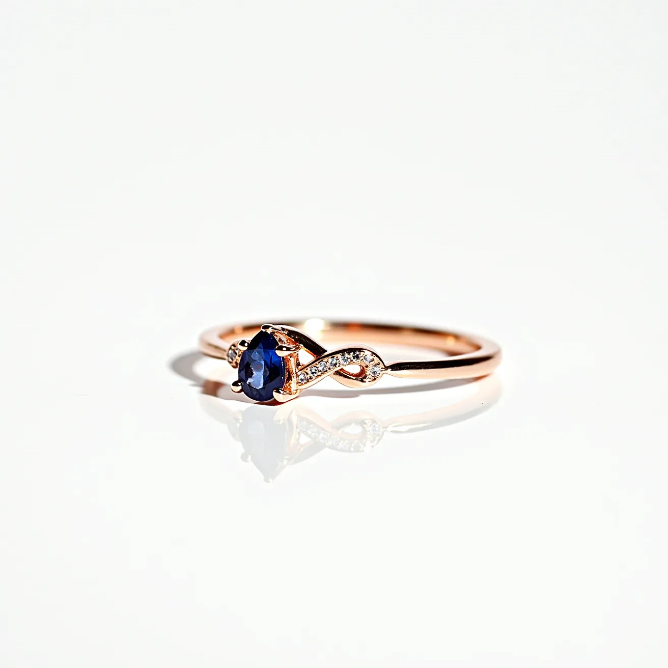 This sapphire ring features a sleek rose gold band, elegantly complementing the deep blue, pear-cut sapphire set at its center. The sapphire is securely held in place by a prong setting, allowing light to highlight its vibrant hue. Adjacent to the sapphire, small round-cut diamonds are intricately set along a curving line, adding a sparkling accent that enhances the ring’s overall elegance. This design combines classic beauty with a modern twist, highlighted by the intricate settings and the contrast between the sapphire and diamonds.