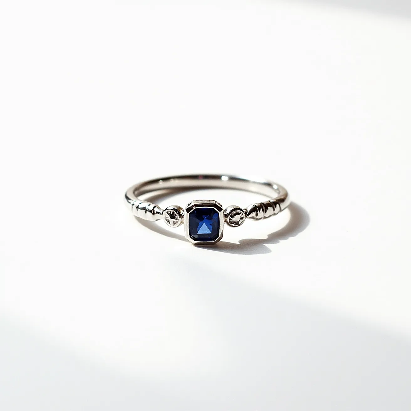 This sapphire ring features a striking square-cut blue sapphire set at its center, embraced by a bezel setting that enhances the stone's elegance and security. The band is likely crafted from a polished white metal, possibly platinum or white gold, contributing to the ring's sophisticated aesthetic. Accentuating the central sapphire are two small metallic bead-like embellishments on the band, adding texture and visual interest. The sleek and minimalistic design of this ring allows the vibrant sapphire to take center stage, making it a timeless piece suitable for various occasions.