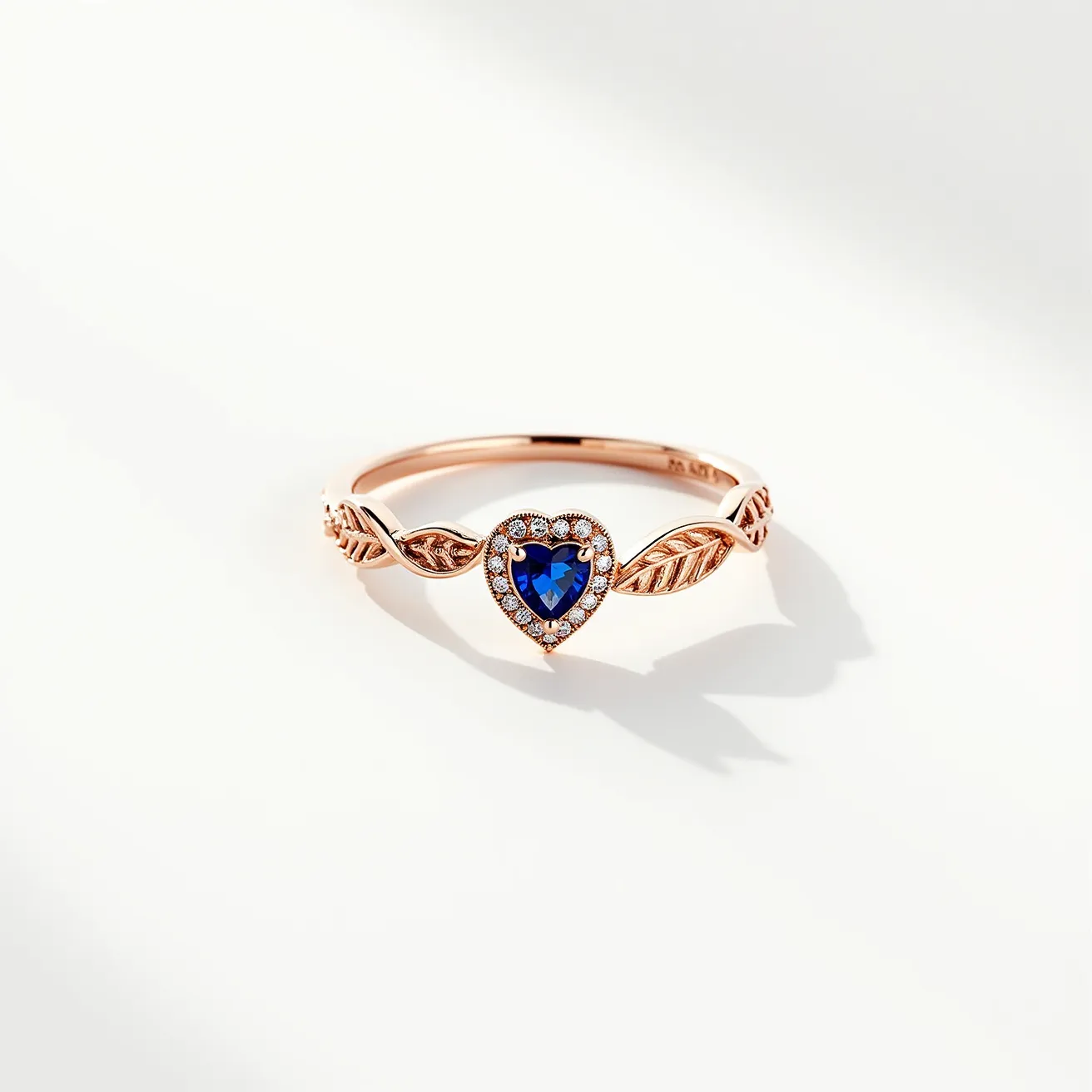 This sapphire ring features a striking heart-shaped sapphire as its central gemstone, surrounded by a halo of smaller, brilliant-cut diamonds. The ring is crafted from a material that appears to be rose gold, enhancing the elegance and warmth of the overall design. The band is adorned with intricate leaf-like engravings on either side of the setting, adding an organic and sophisticated touch. The setting securely holds the central sapphire, with the diamonds enhancing the brilliance and color of the main stone. The ring’s design seamlessly combines classic elements with a modern twist, making it both timeless and unique.