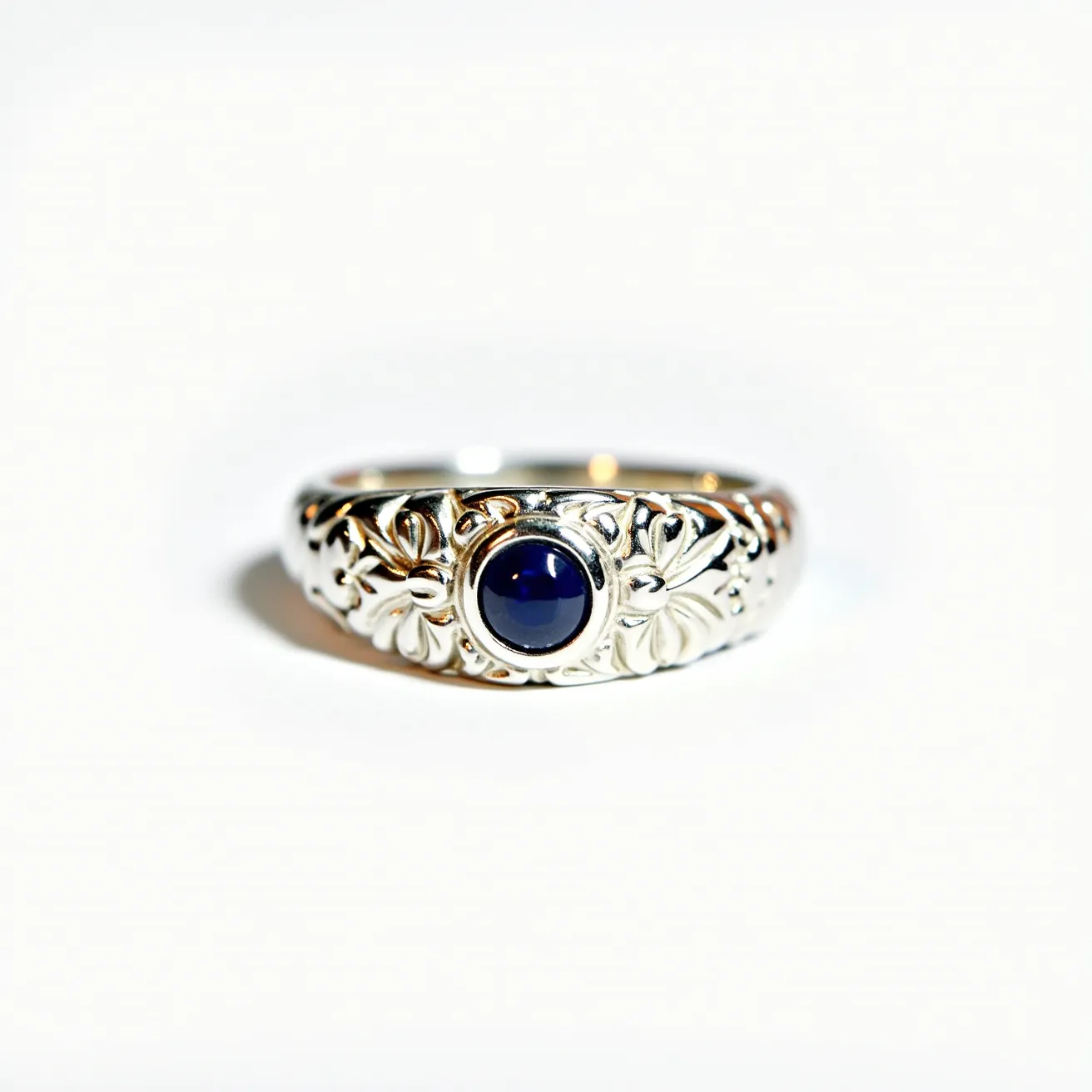 This signet ring features an intricate floral design crafted in what appears to be silver or a similarly lustrous metal. At the center of the ring is a polished, dark blue gemstone, possibly a sapphire, presented in a round cabochon cut which allows the stone to sit smoothly and prominently. The stone is set in a bezel setting, securely encasing the gem within the metal. The detailed etching and high-quality craftsmanship contribute to the ring’s elegant and timeless appearance, making it a piece that stands out both in design and function.