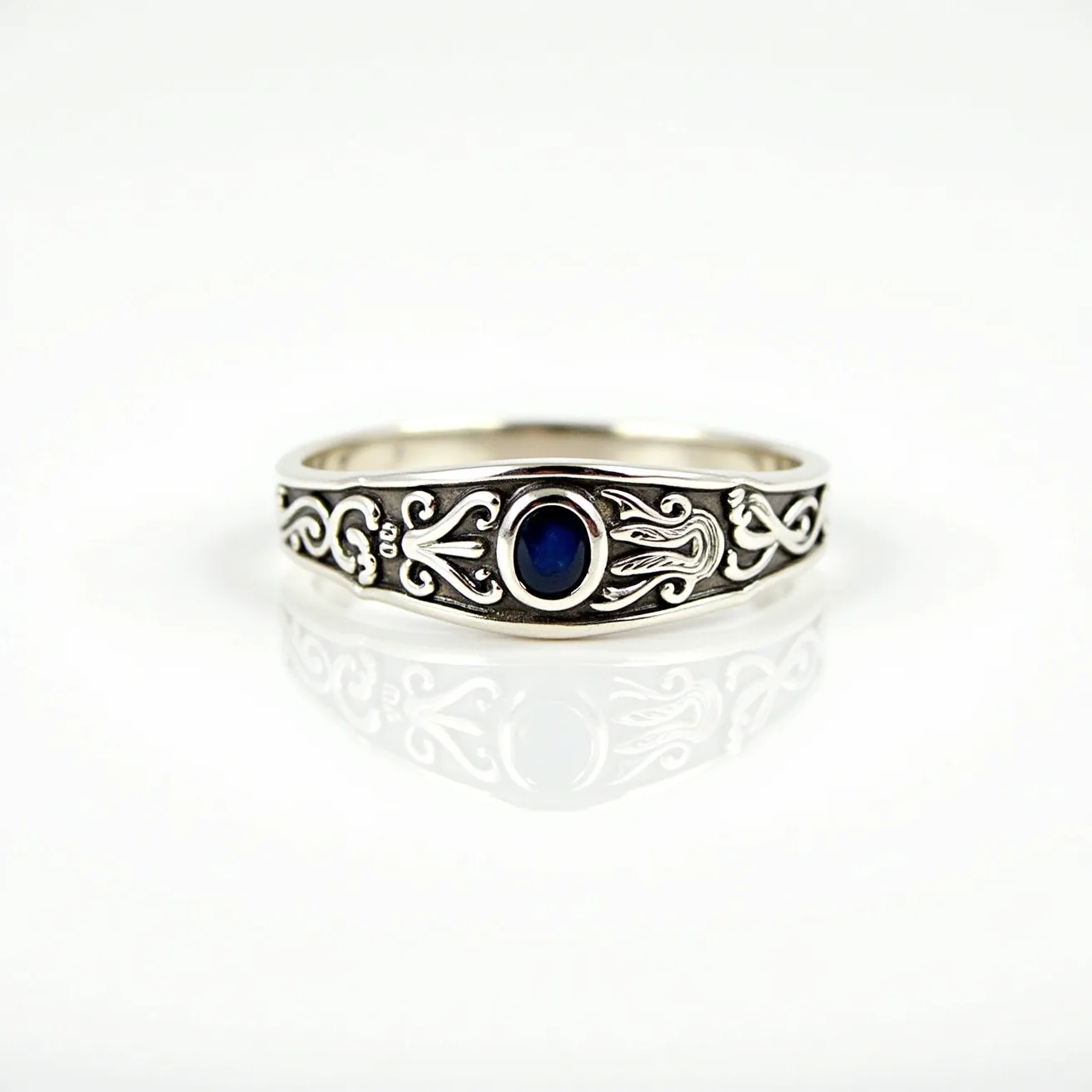 This signet ring features intricate silver craftsmanship with detailed engravings along the band. At its center is a small, round, deep blue sapphire, expertly set in a bezel setting that ensures a secure fit. The silver metal complements the rich blue of the stone, highlighting its elegance and refinement. The band is smooth and unified, providing a seamless and polished look without additional clasps or attachments.