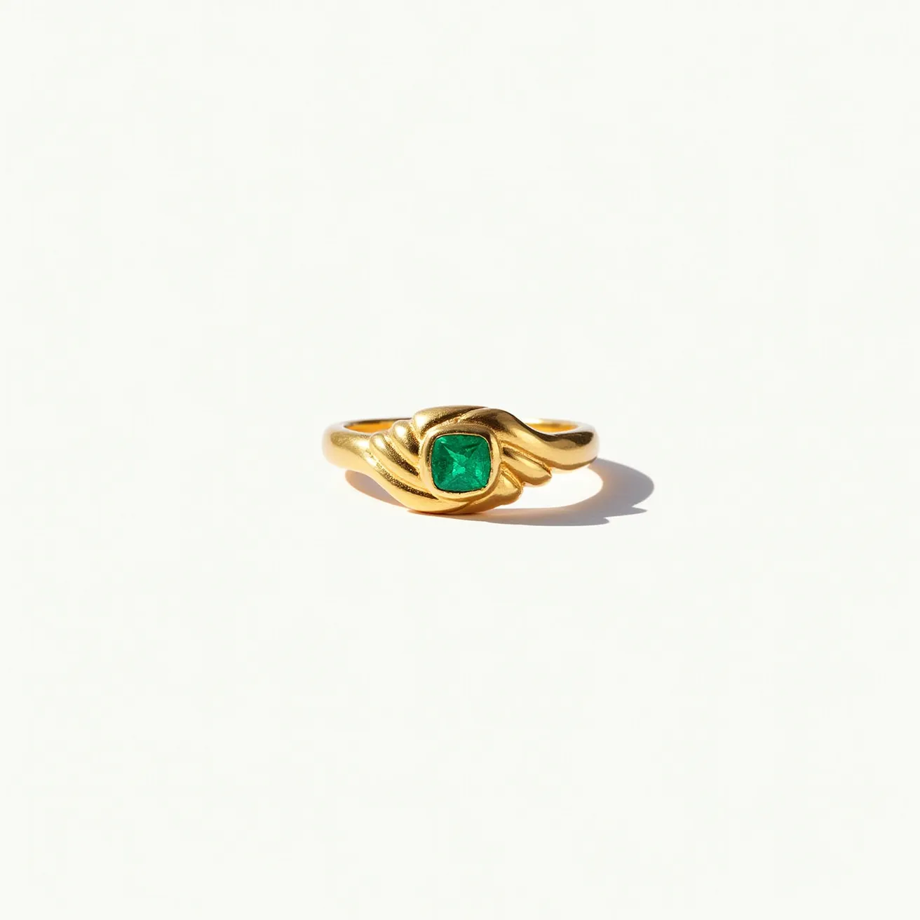 This signet ring features a beautifully set, square-cut green gemstone, likely an emerald, at its center. The gem is encased in a smooth gold band that elegantly swirls around it, enhancing its prominence. The gold band showcases a polished finish, adding to the ring’s sophisticated and timeless appeal. The gemstone is securely mounted in a bezel setting, which provides both protection and a seamless look. The ring’s design is characterized by its graceful, flowing lines, creating an impression of classic elegance paired with modern simplicity.