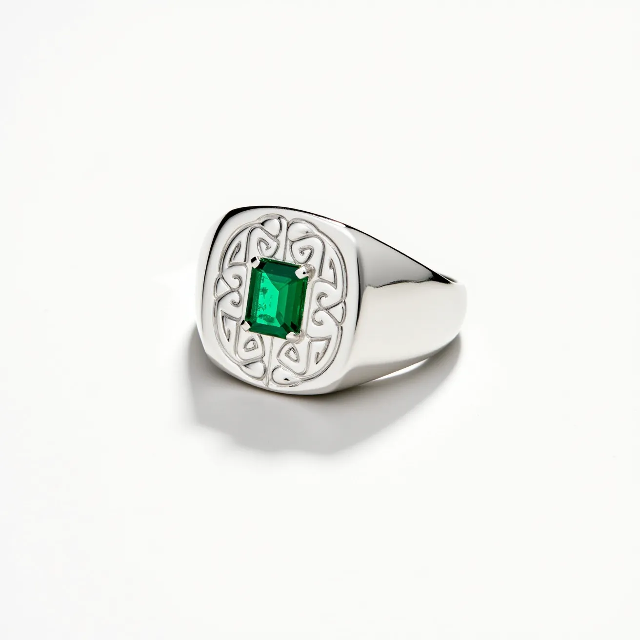 This signet ring features a polished metal band that is likely fashioned from silver or white gold. It is adorned with an emerald-cut green gemstone securely mounted in a prong setting. Surrounding the stone is an intricately designed motif that adds elegance and a touch of classic detailing to the piece. The stone’s vivid green hue contrasts beautifully with the smooth, lustrous metal of the band, creating a striking appearance. The ring is crafted to fit comfortably with a slightly tapered shank that supports the detailed face, making it both a stylish and enduring accessory.