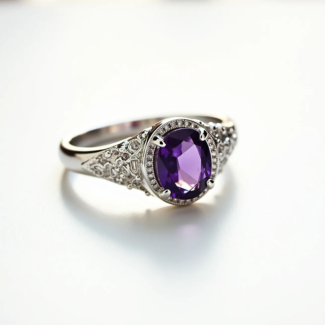 This signet ring features an elegant design crafted from silver or white gold, which serves as the band and the intricate base for the ring. At its center, a striking oval-cut purple gemstone, likely an amethyst, is set, exhibiting a rich and vibrant hue. The gemstone is secured with a classic four-prong setting, enhancing its prominence and securing it firmly in place. Surrounding the central gem, detailed engravings and small accent stones complement the design, adding to the ring's sophisticated and ornate appearance. The band showcases intricate filigree work which adds texture and complexity to the overall aesthetic of the piece.