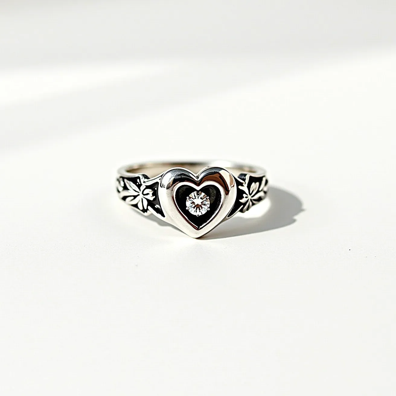 This silver heart ring features a polished heart-shaped centerpiece made from silver, adorned with intricate floral detailing on the band. At the center of the heart is a round-cut gemstone, likely a diamond, set in a bezel or prong setting, adding a touch of sparkle to the piece. The ring is crafted to showcase an elegant design without the need for any clasps or attachments, maintaining a classic and seamless appearance.