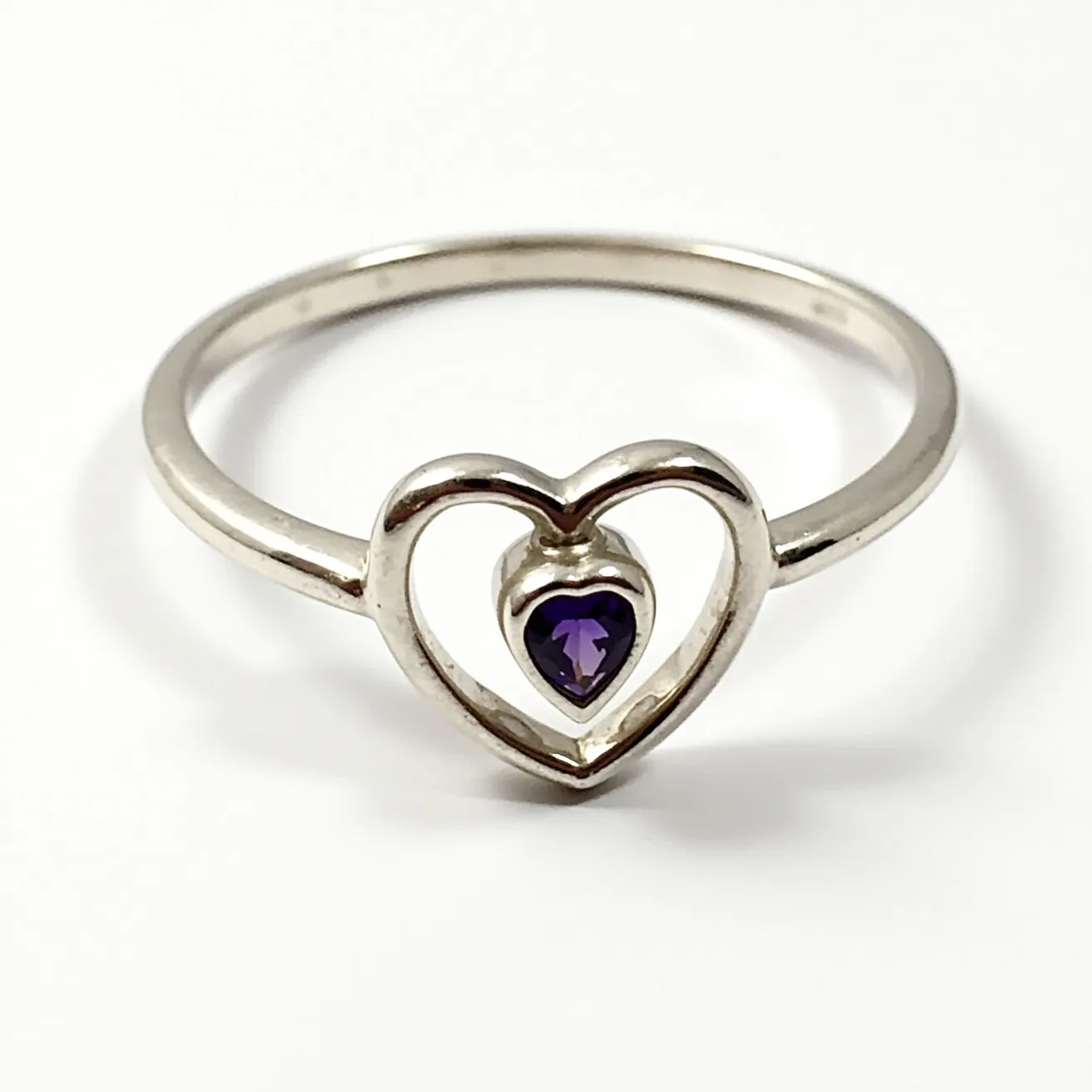 This silver heart ring features a delicate and polished metal band that supports a central heart motif. Within the open heart design, there is a single heart-shaped gemstone, likely an amethyst, showcasing a deep purple hue. The gemstone is bezel-set, adding a secure and smooth finish to its presentation. The ring’s minimalist design highlights the gemstone and heart shape, creating a charming and elegant piece of jewelry.