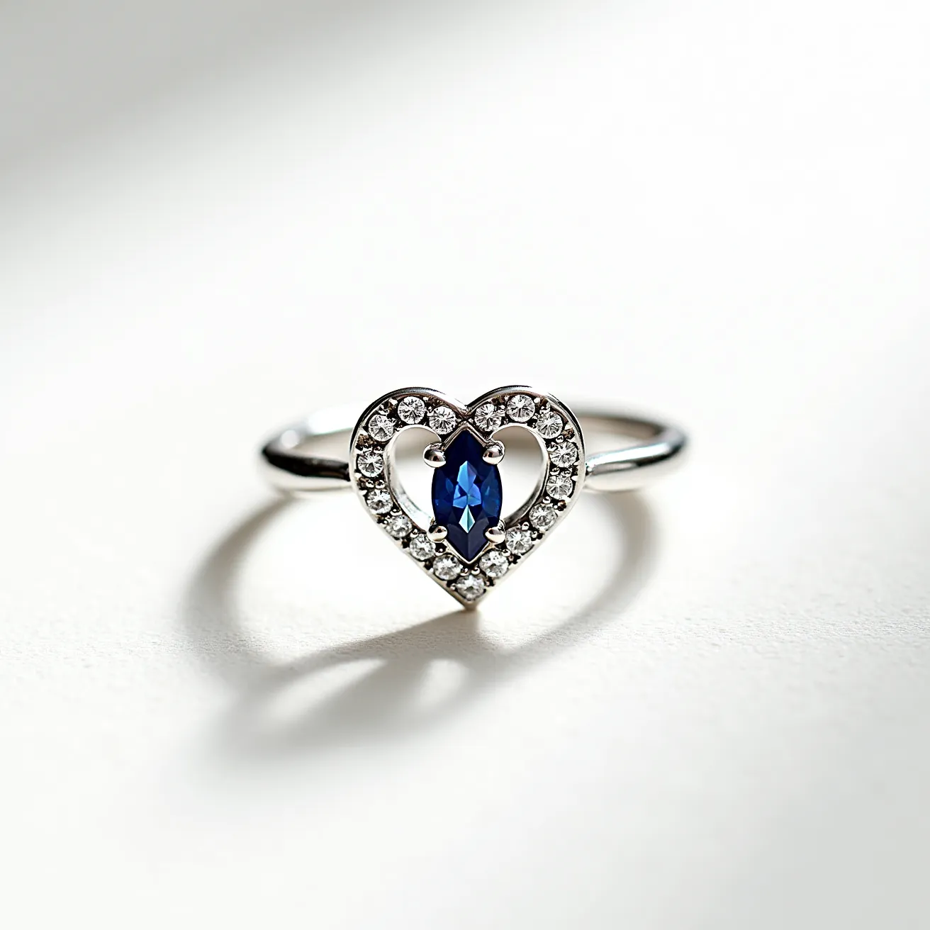 This silver promise ring features a delicate heart-shaped design encrusted with small, clear gemstones studded in a pave setting, creating a sparkling border. At the center of the heart lies a marquise-cut blue stone set with prongs, drawing attention with its striking color and shape. The band is simple and polished, seamlessly supporting the intricate heart centerpiece. The overall craftsmanship combines elegance and symbolism, making it a meaningful piece of jewelry.